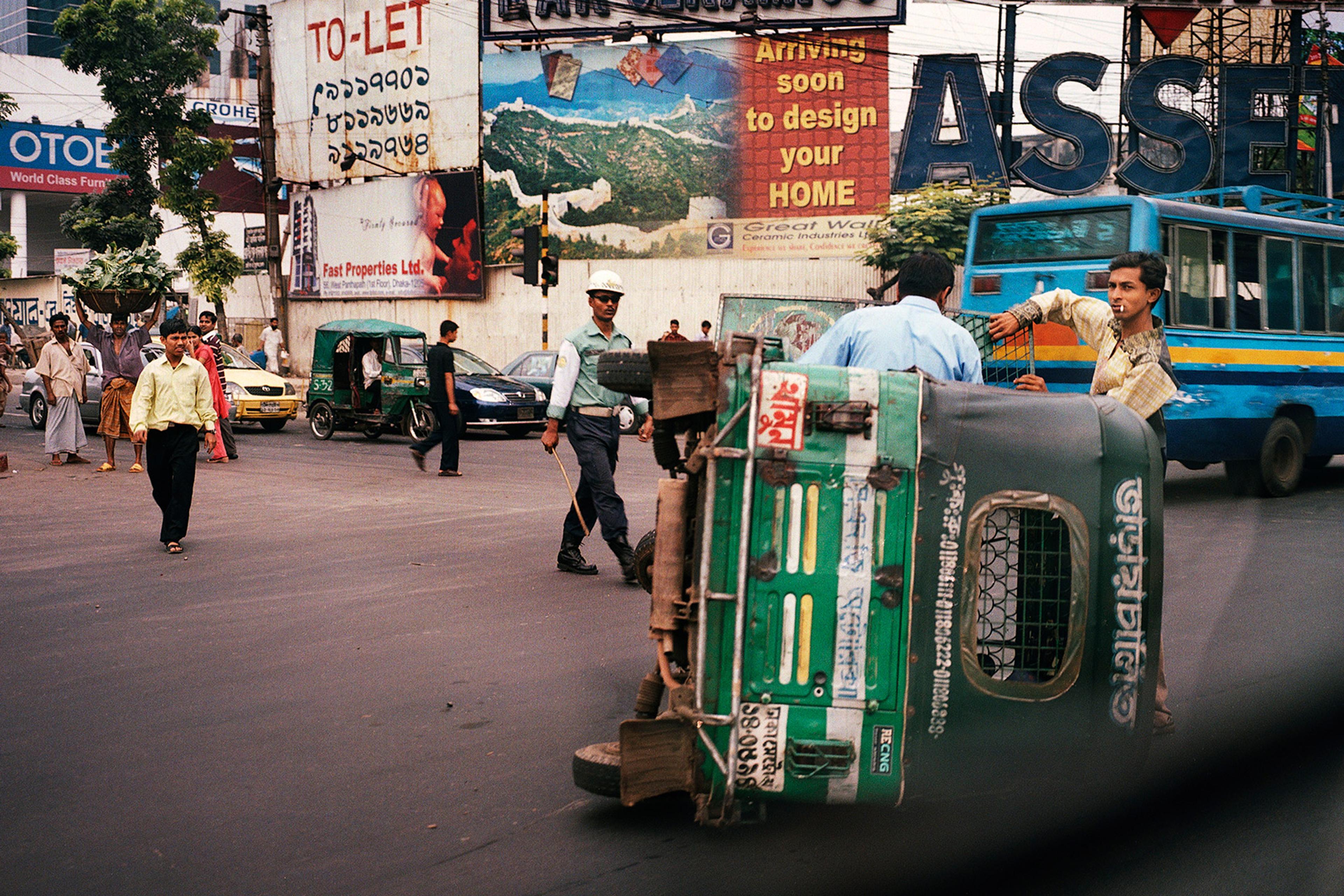 A busy street with billboards, people walking and police officers beside an overturned green auto-rickshaw, with a blue bus and other vehicles passing by.