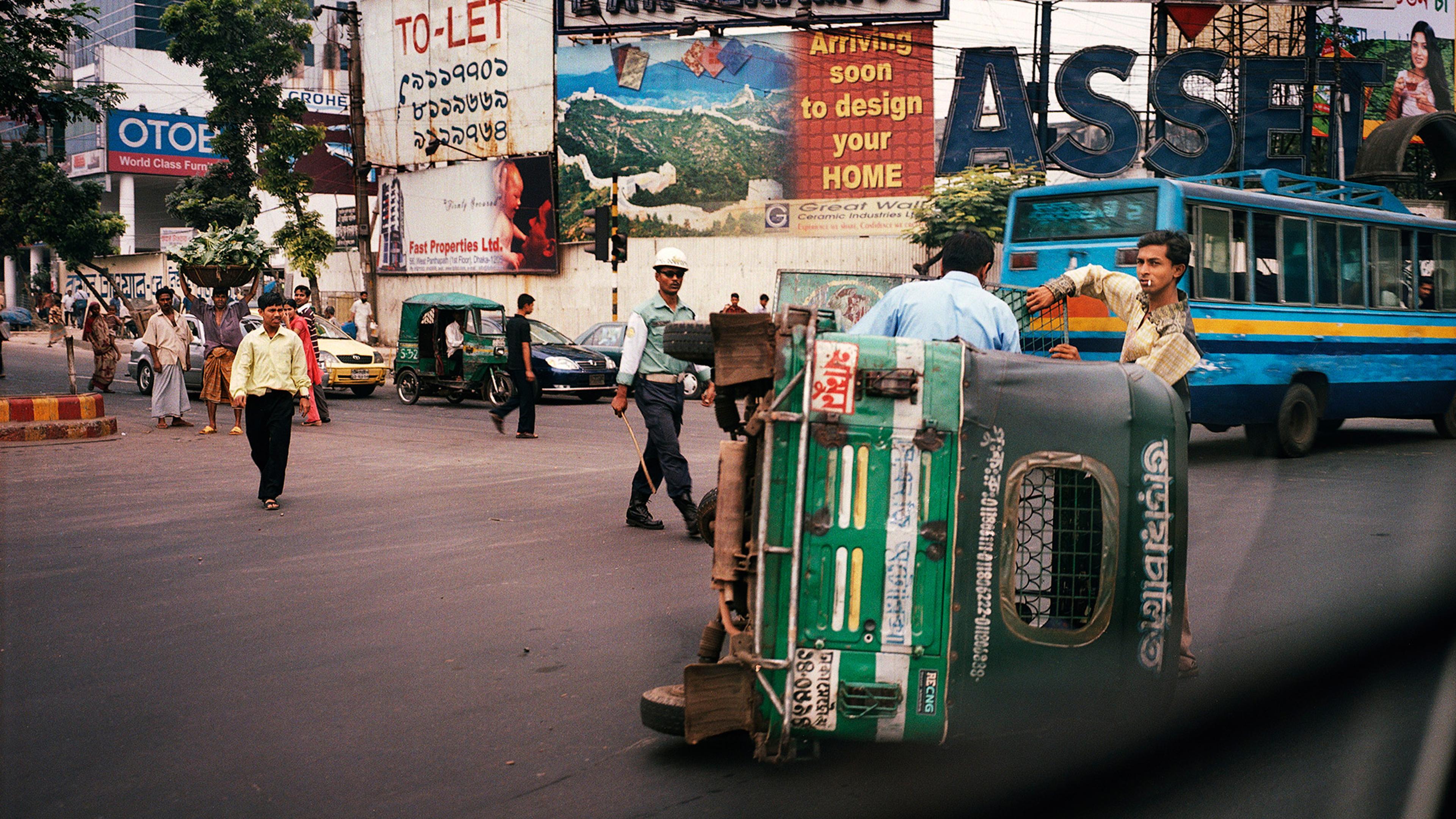 A busy street with billboards, people walking and police officers beside an overturned green auto-rickshaw, with a blue bus and other vehicles passing by.