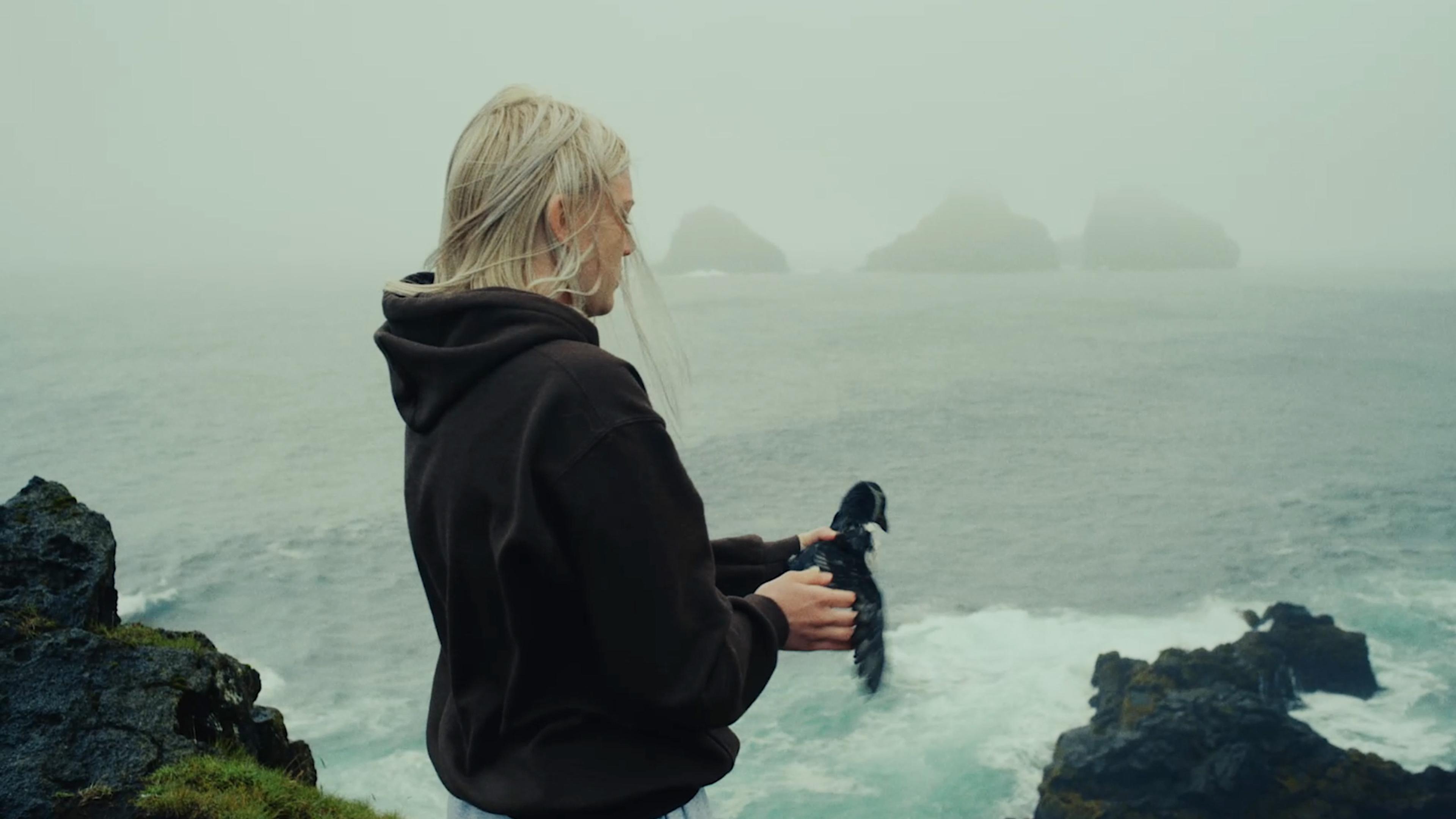 A person with light blonde hair, wearing a dark hoodie, stands on a cliff by the sea, holding a bird. The sea is rough with waves crashing against the rocks, and several rock formations are visible in the misty background.