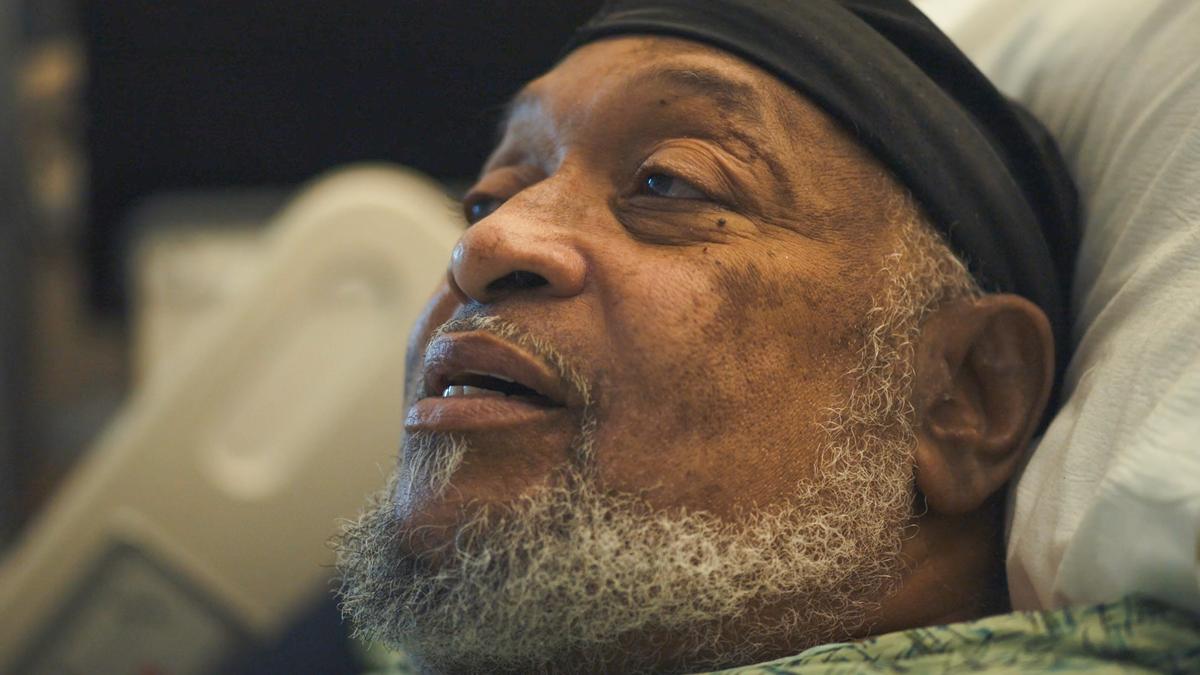 Close-up photo of an elderly man with a beard wearing a black cap, lying in a hospital bed, looking content.