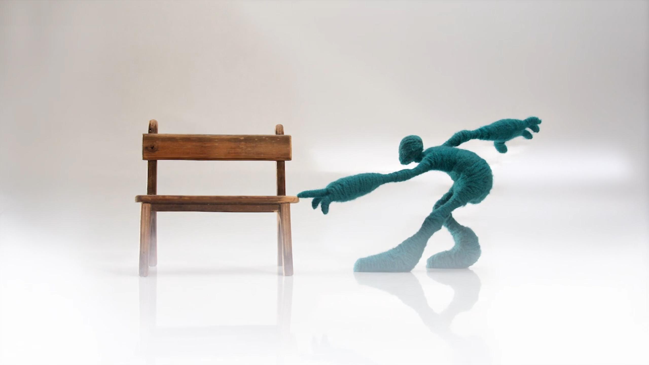 Sculpture of a human-like figure made of teal wool reaching towards a wooden chair on a light background.