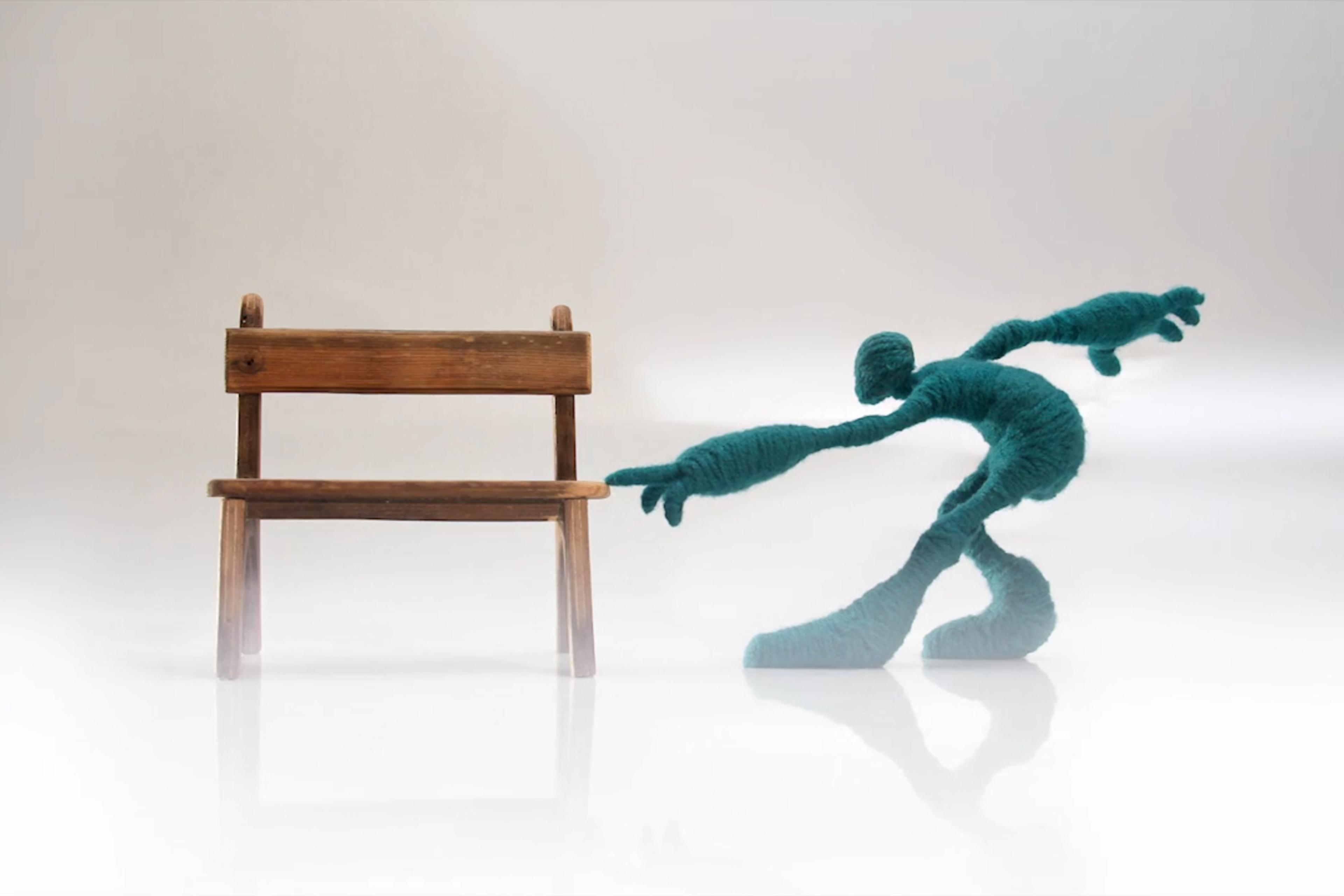 Sculpture of a human-like figure made of teal wool reaching towards a wooden chair on a light background.