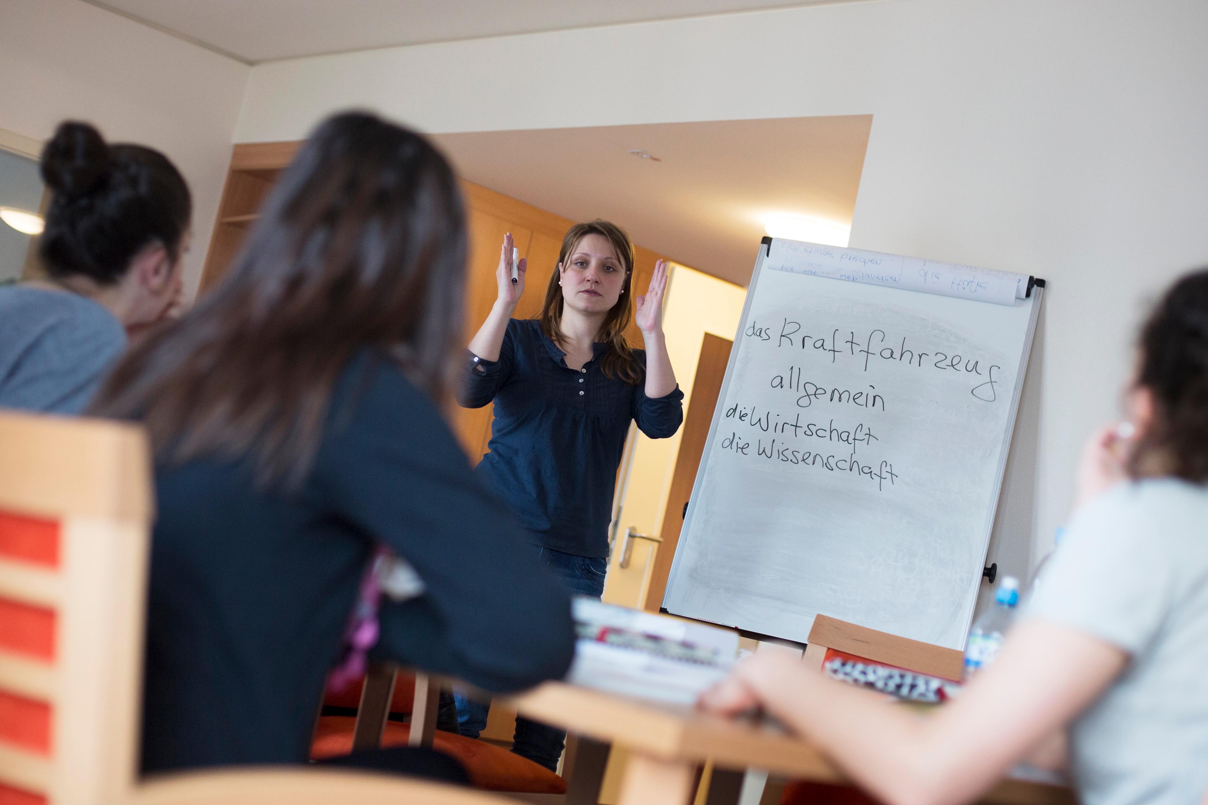 Photo of a woman teaching a class with a whiteboard displaying German words, students seated in foreground.