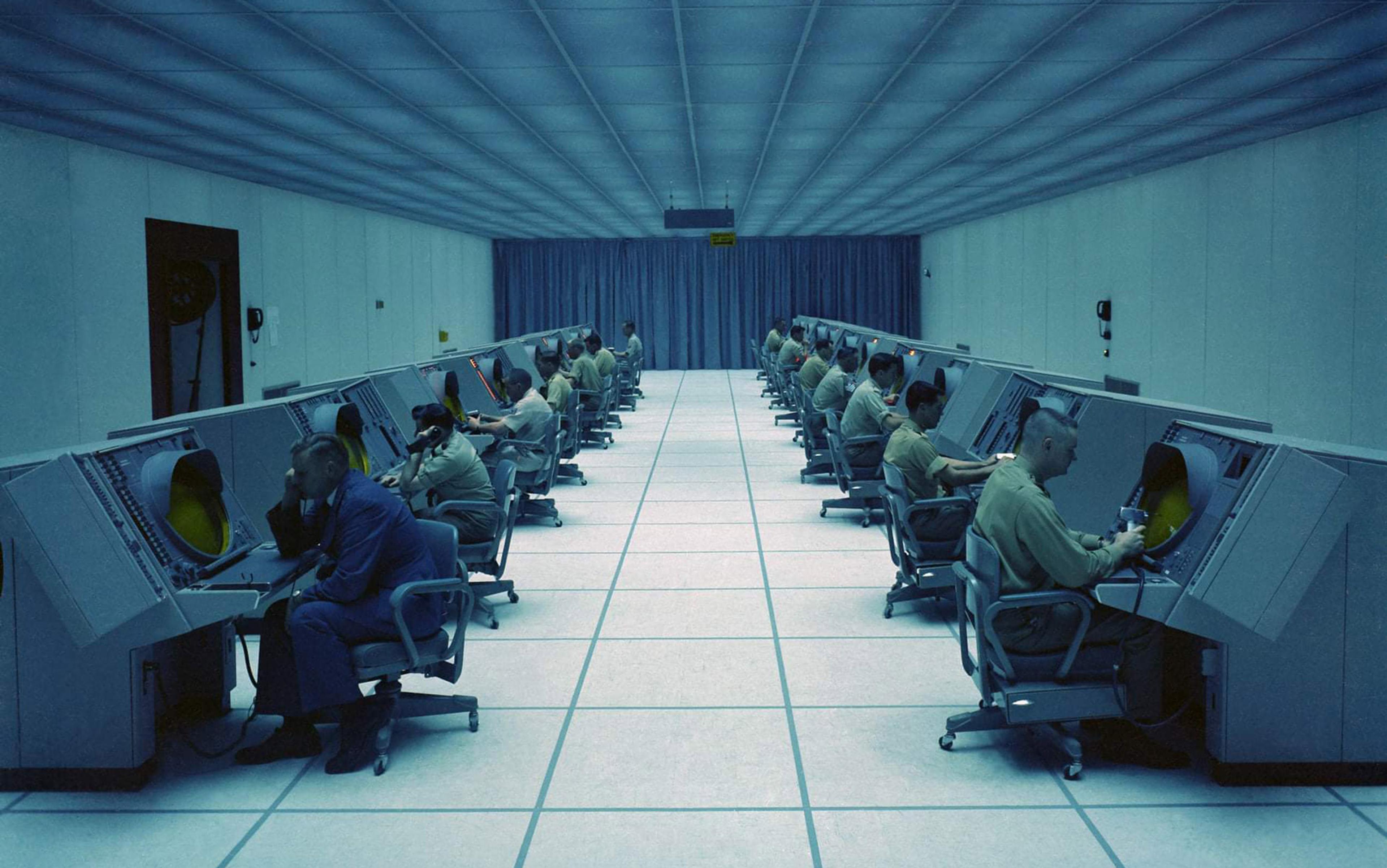 A large control room with people sitting at consoles in two rows, facing away from each other, under blue lighting.