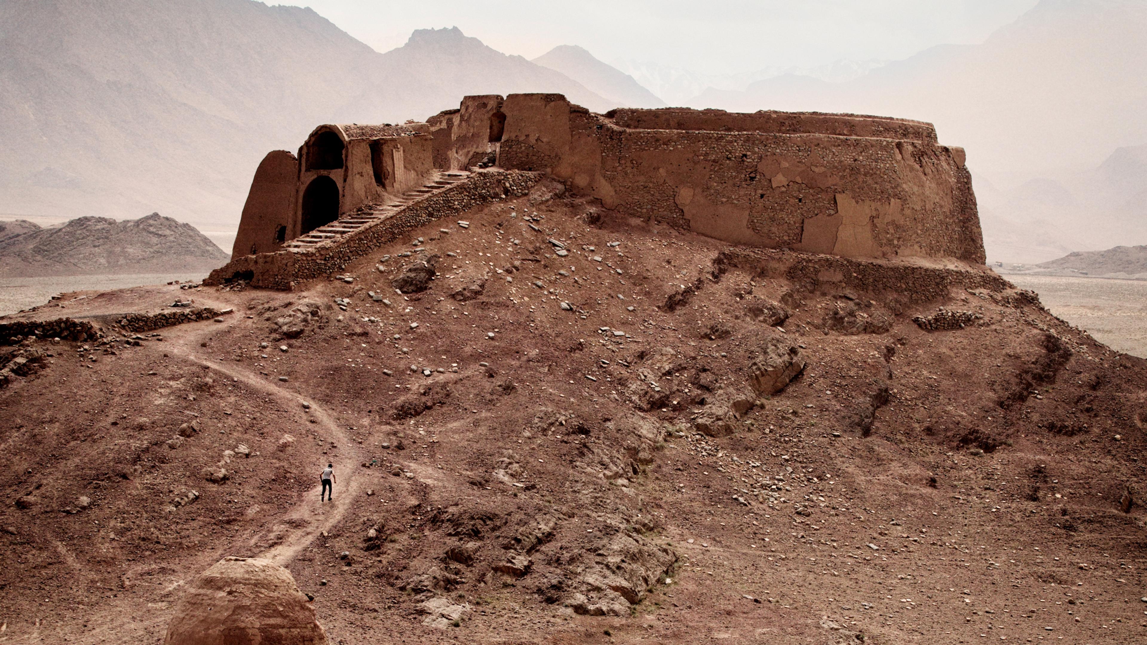 Photo of ancient ruins on a rocky hill with a cloudy sky and distant mountains in the background.