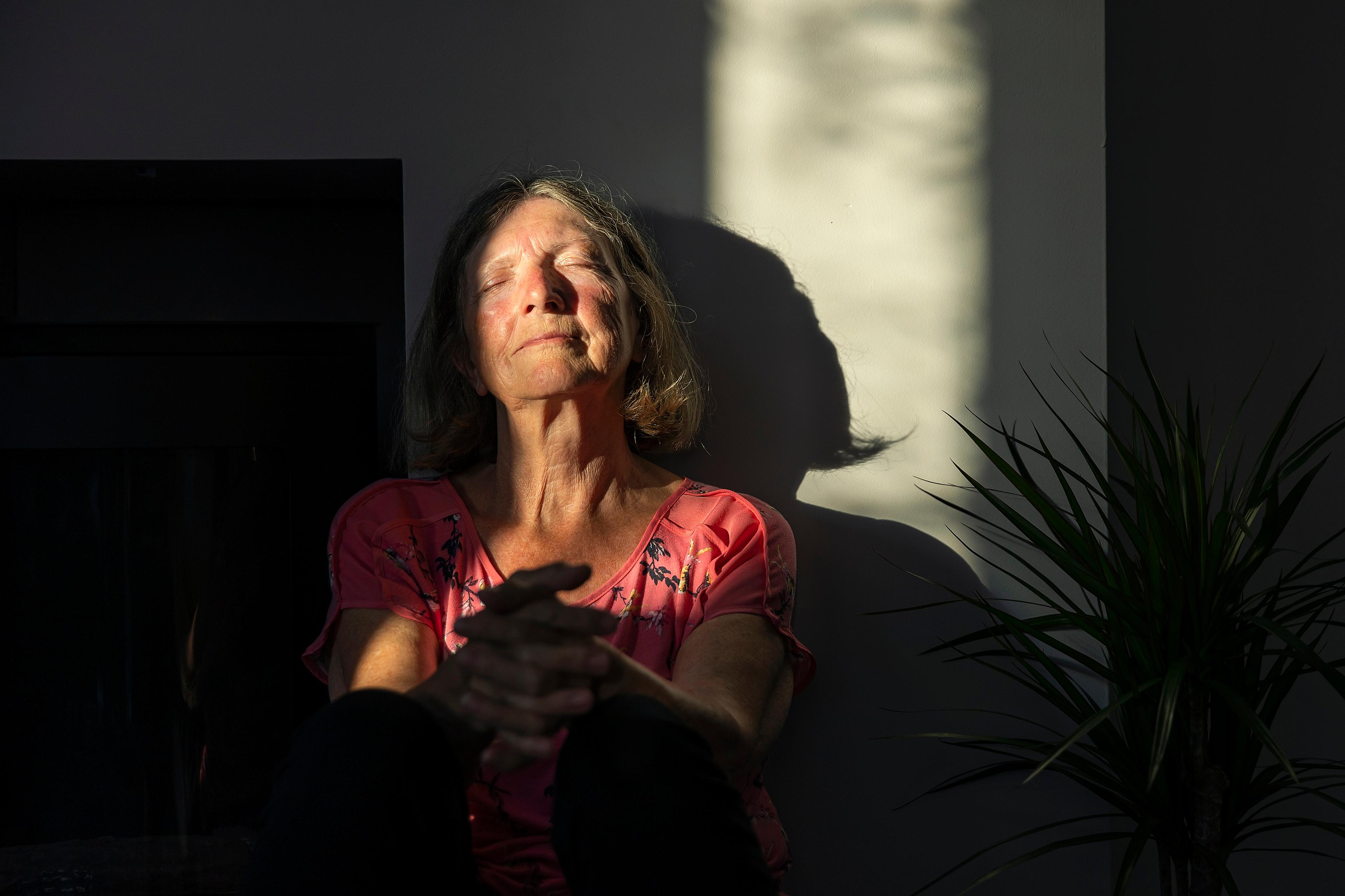 Photo of a woman in a pink top sitting indoors with eyes closed. Sunlight and shadows on the wall, plant on the right.
