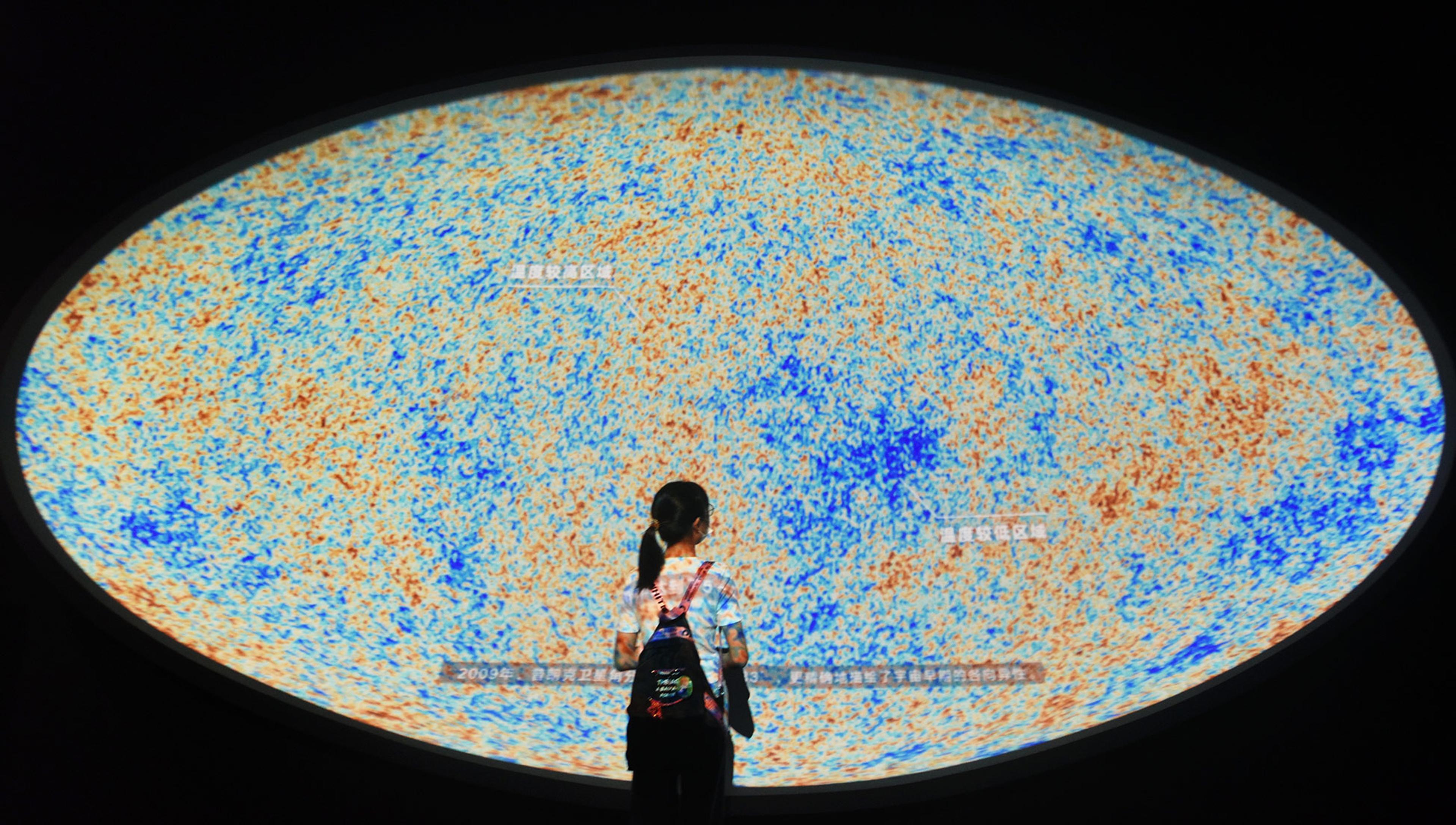 A young girl, seen from the back, in a museum in China stands before a huge, oval multicoloured image of ‘fossil’ microwave radiation