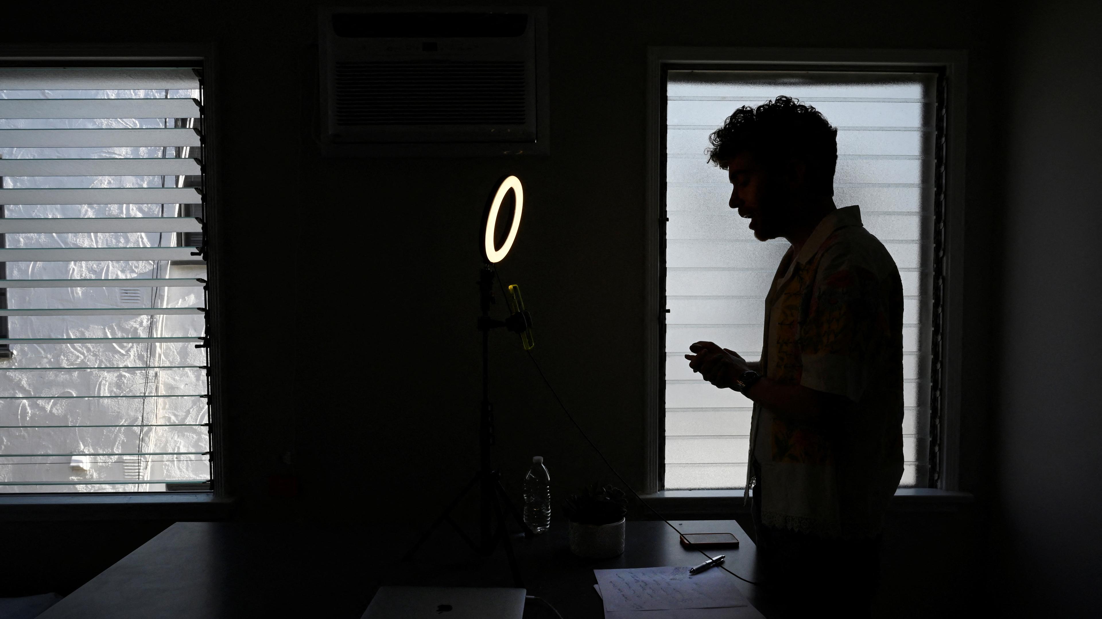 Silhouette of a man by a window in a dim room with a ring light laptop and papers on the table, photo.