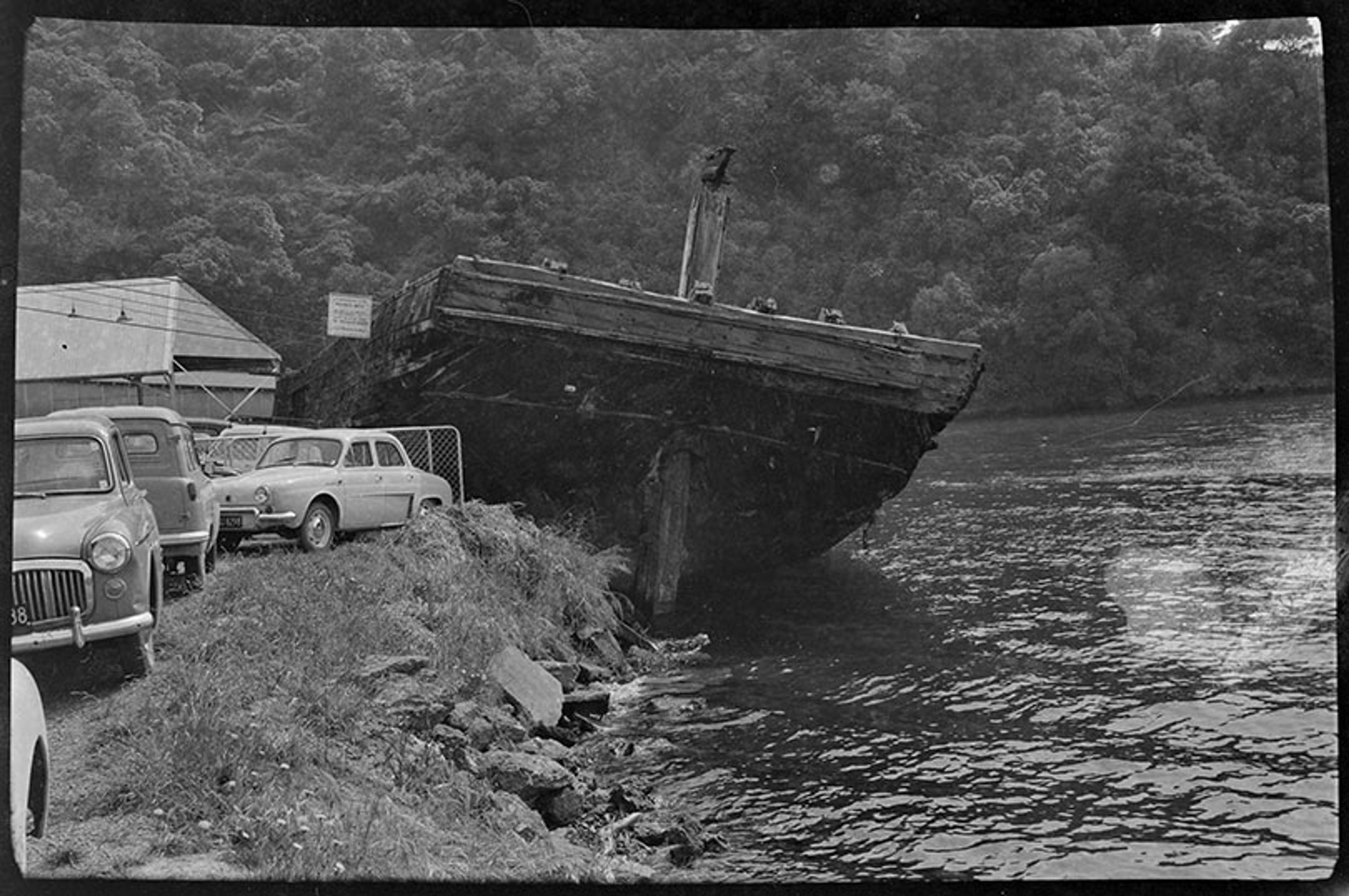Black-and-white photo of a large, old boat beached on a lakeshore near cars and a shed, with a forested hill in the background.