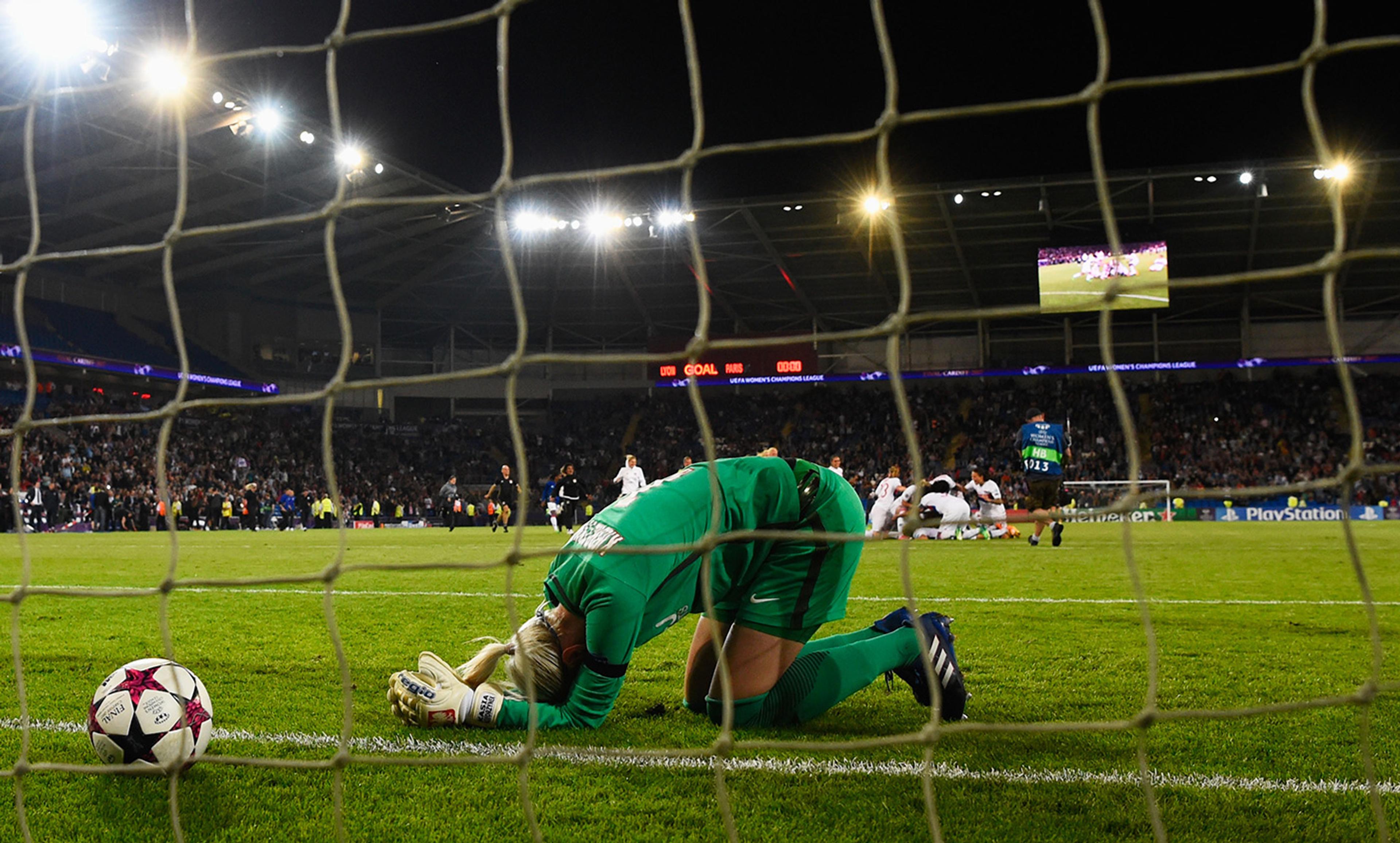 A football goalkeeper kneeling in distress with her face on the grass as the opposing team celebrates a goal under stadium lights.