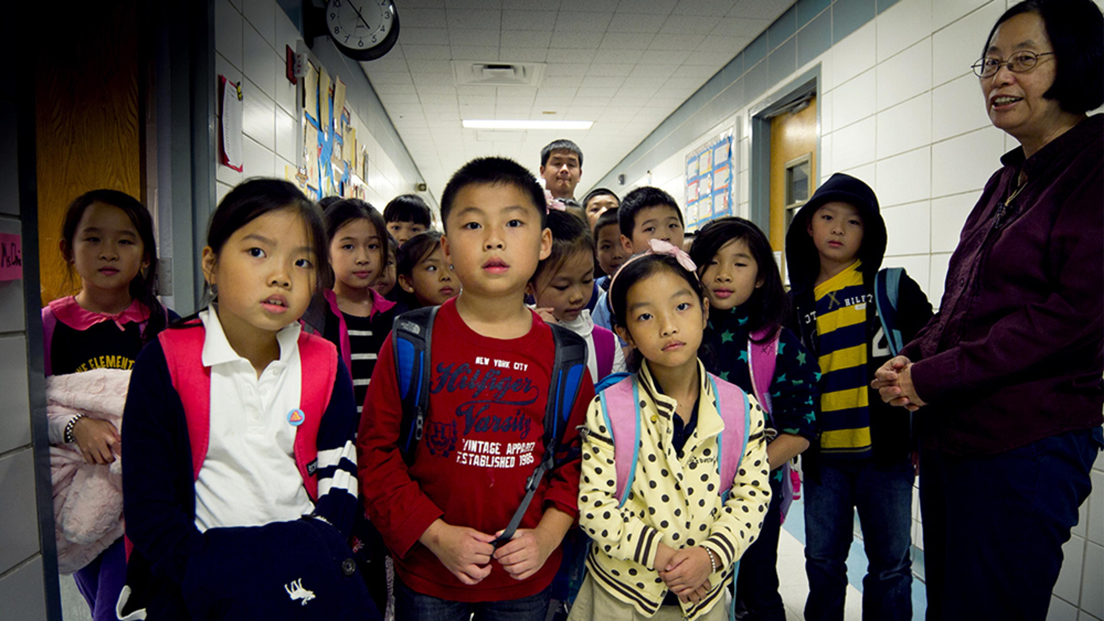 A group of school children with backpacks standing in a corridor with two adults, a clock on the wall above.