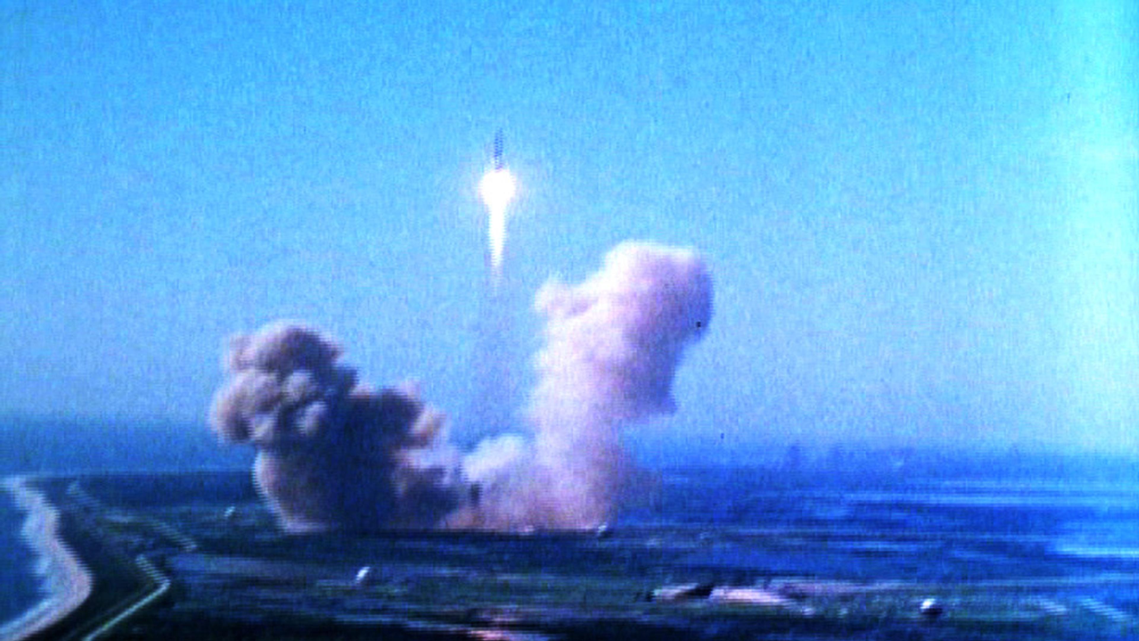 A rocket launch with smoke and flames at the base against a blue sky, captured from a distance at ground level.