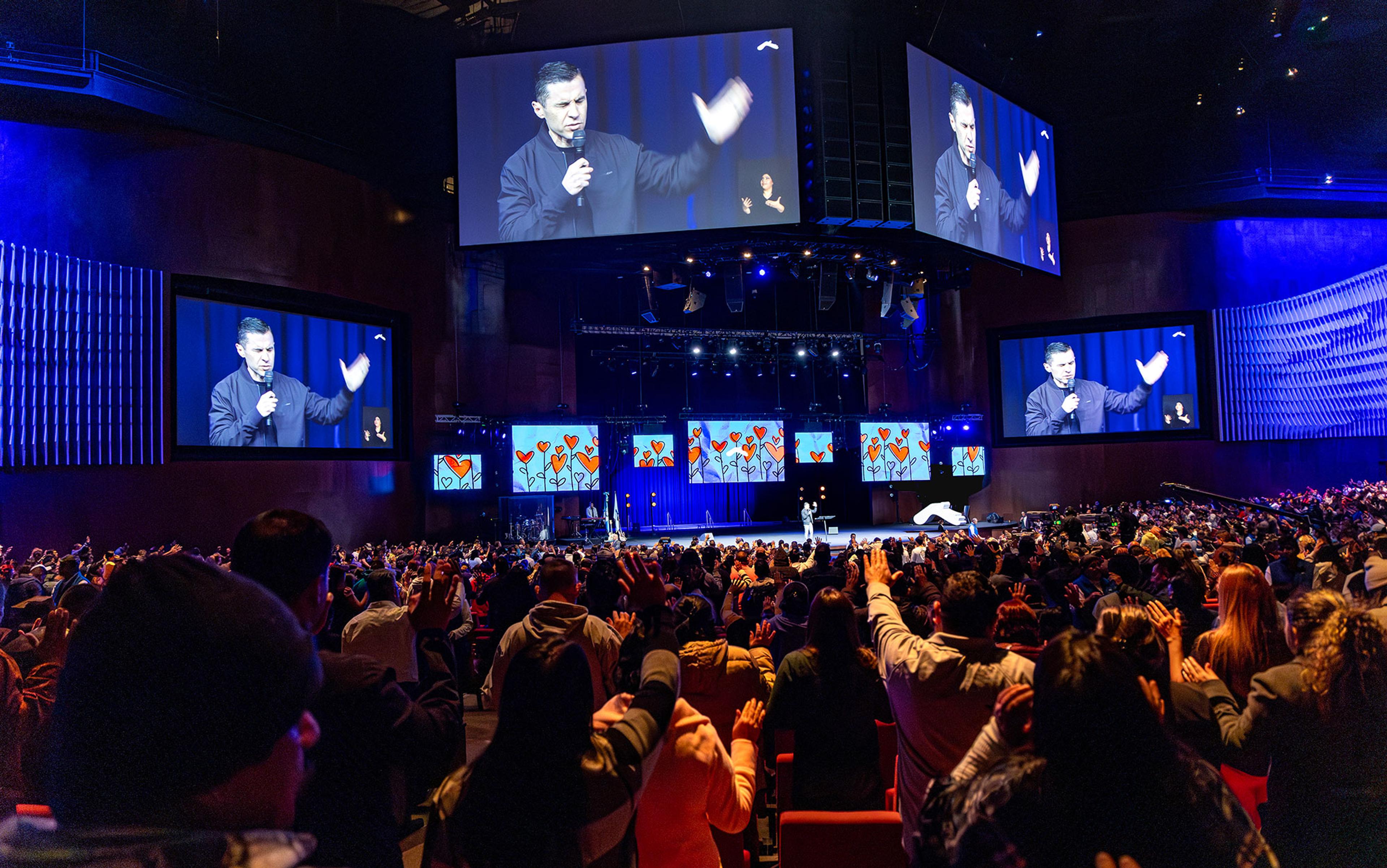 A speaker on stage addressing a large audience in an indoor venue with screens displaying his image.