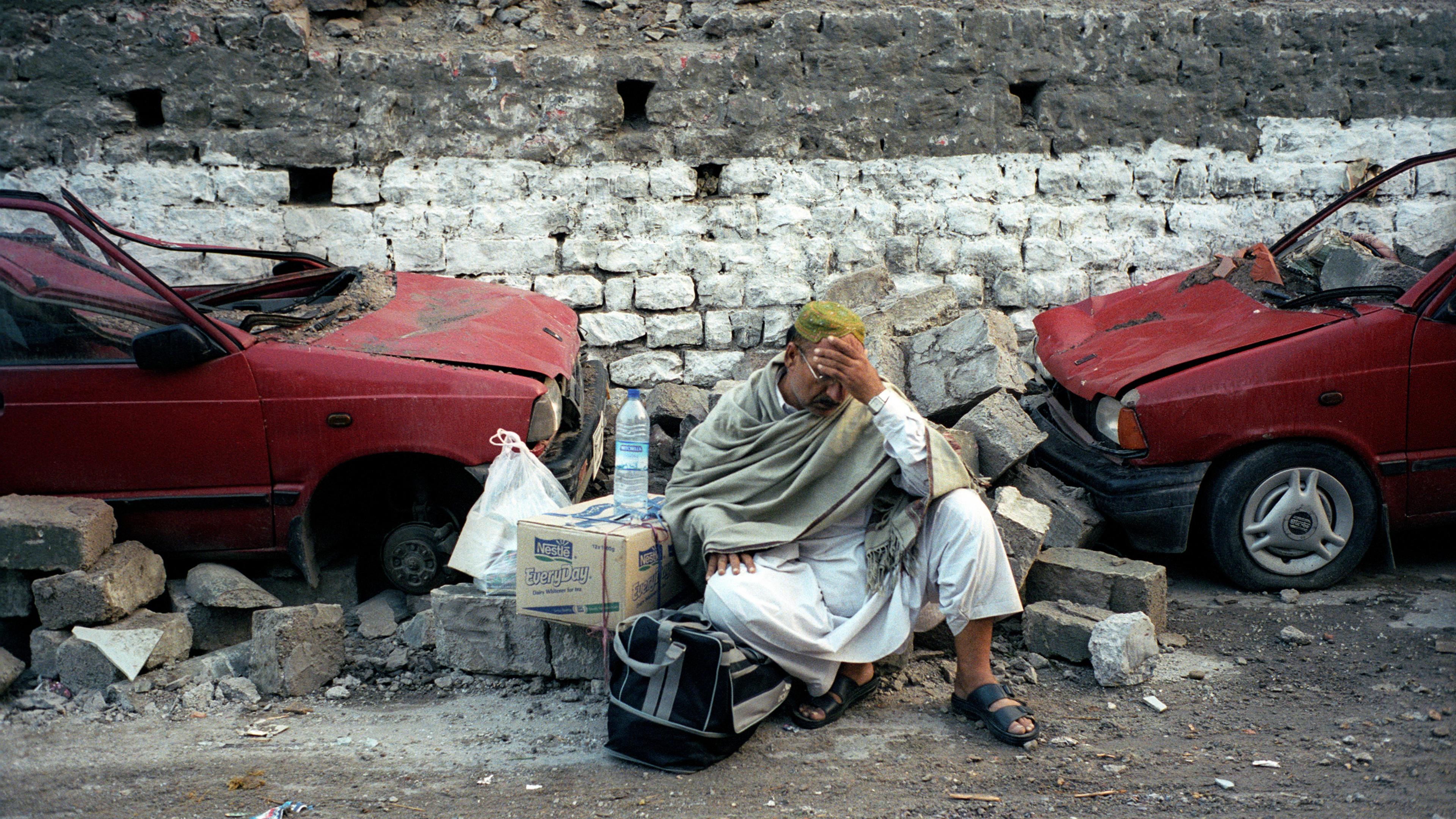 A man wearing a robe sitting amidst rubble between two damaged red cars. He holds his head and has boxes and bags beside him.