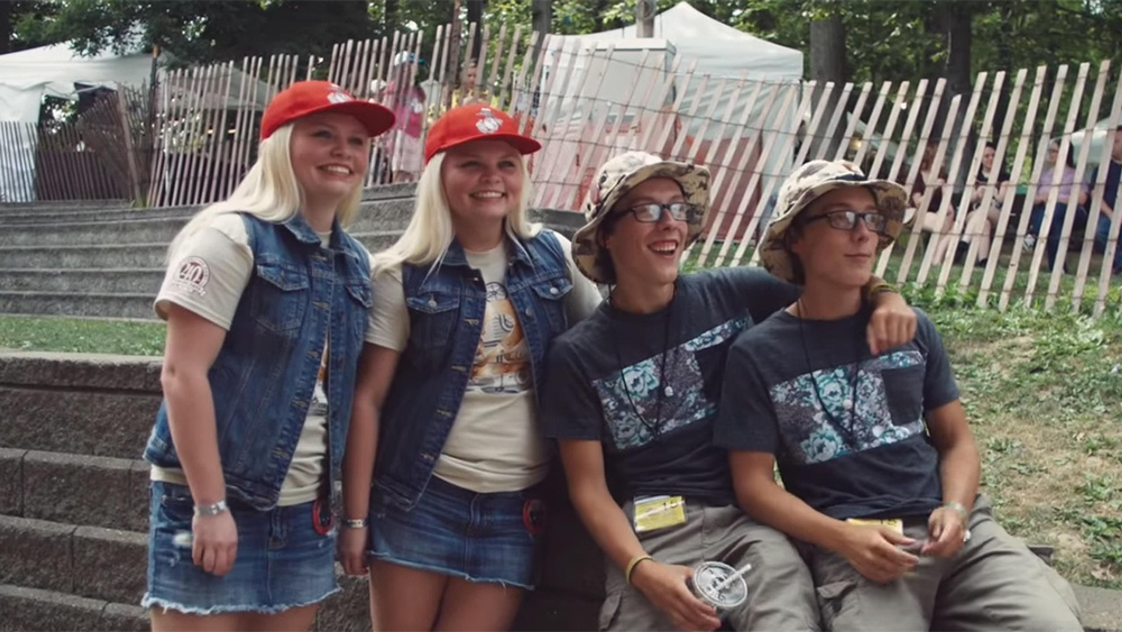 Two sets of smiling young twins outdoors, one set is blonde and female, in denim vests and red caps, the other is male, in glasses, grey shirts and bucket hats, sitting on steps.