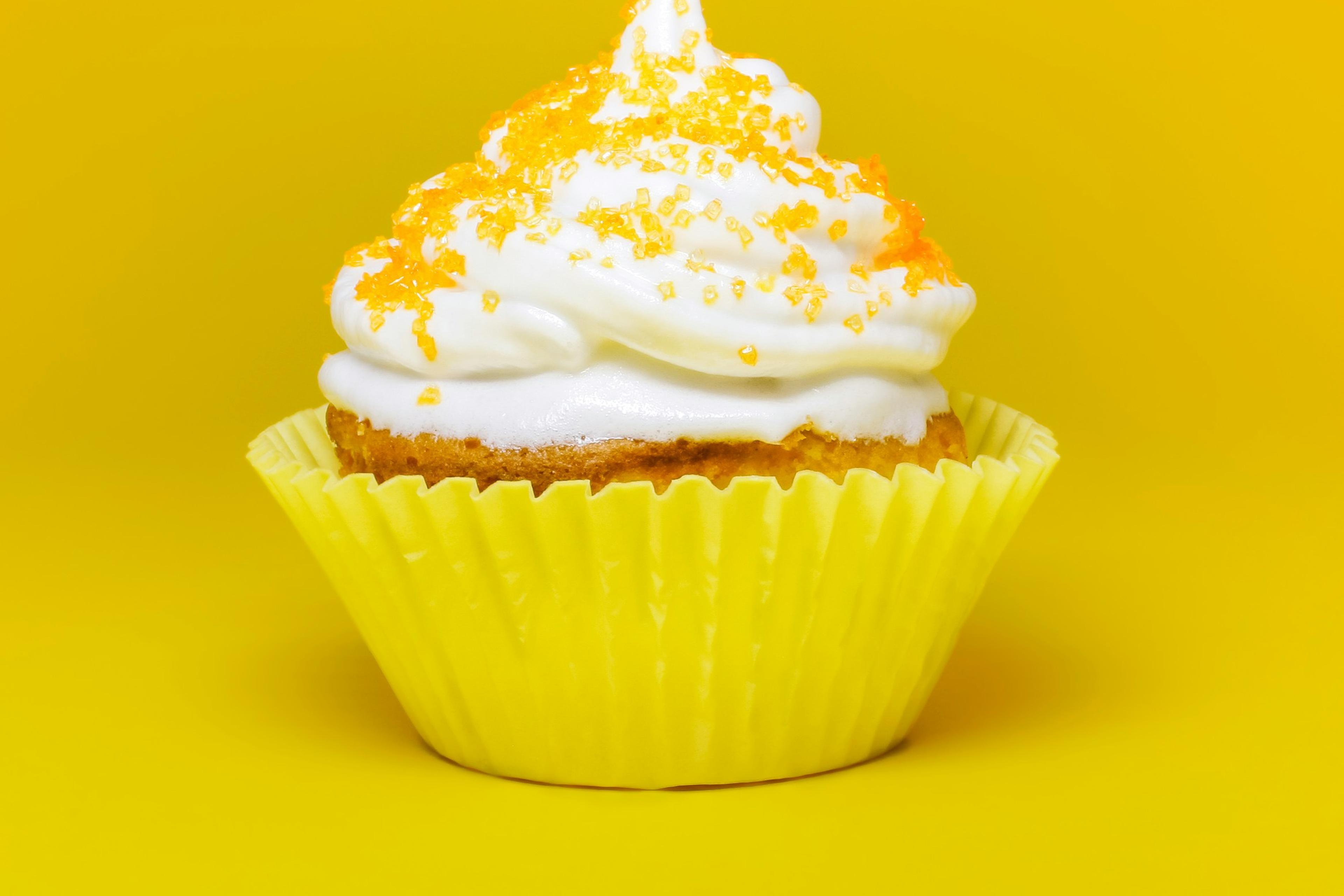 Photo of a cupcake with white icing and orange sprinkles in a yellow wrapper against a bright yellow background.