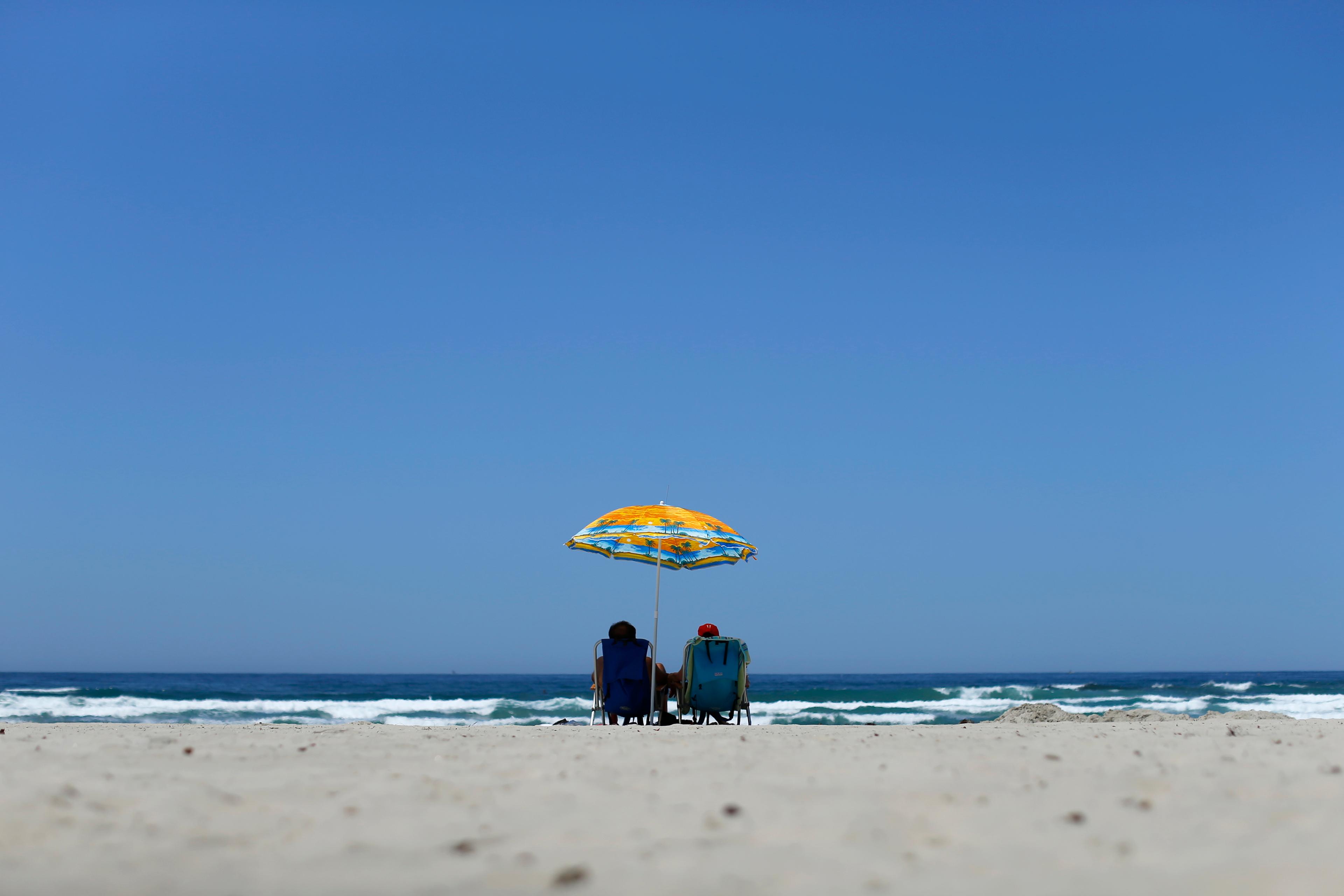 Photo of two people sitting on a beach under a colourful umbrella facing the sea with a clear blue sky.