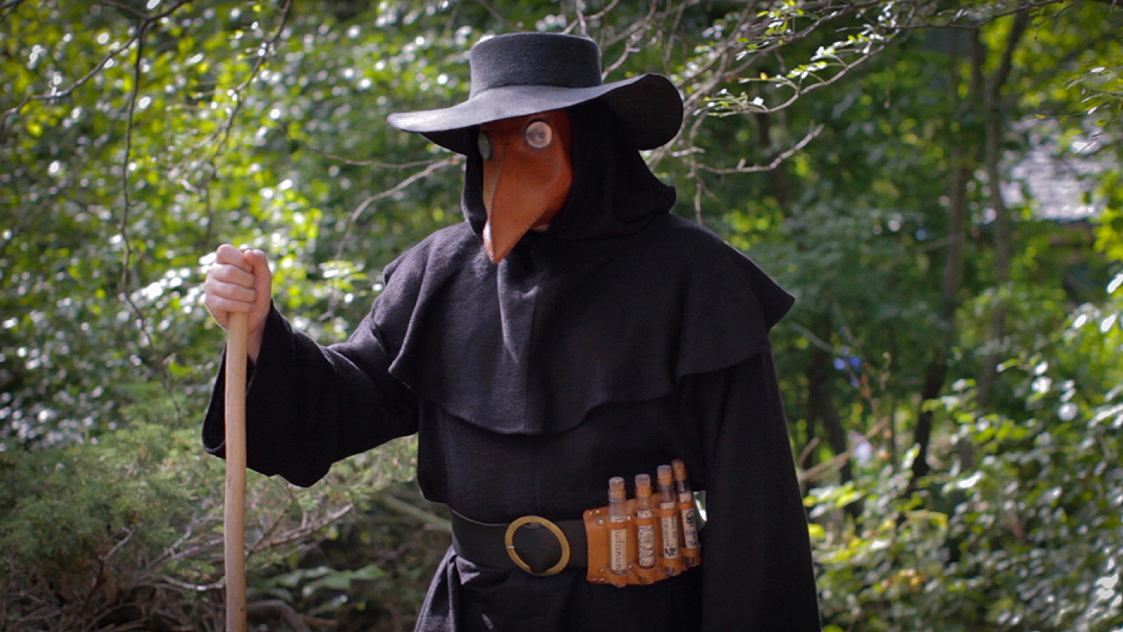 A person in a plague doctor costume with a beaked mask, black cloak and hat, holding a staff in a wooded area during daylight.