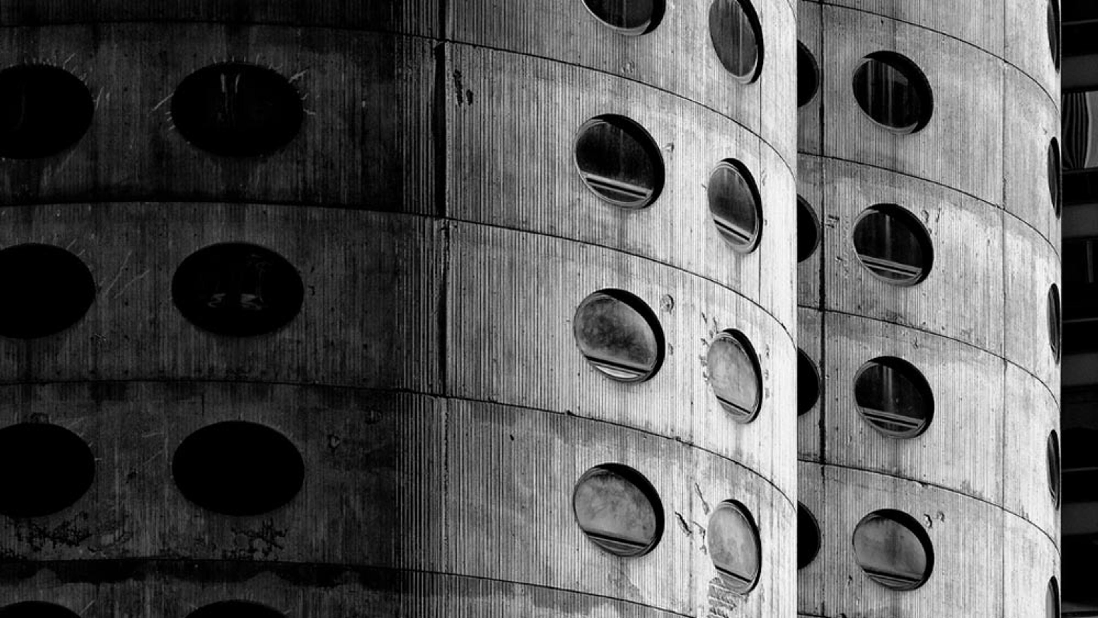 Black and white photo of a cylindrical, brutalist building facade with round windows arranged in a grid-like pattern.
