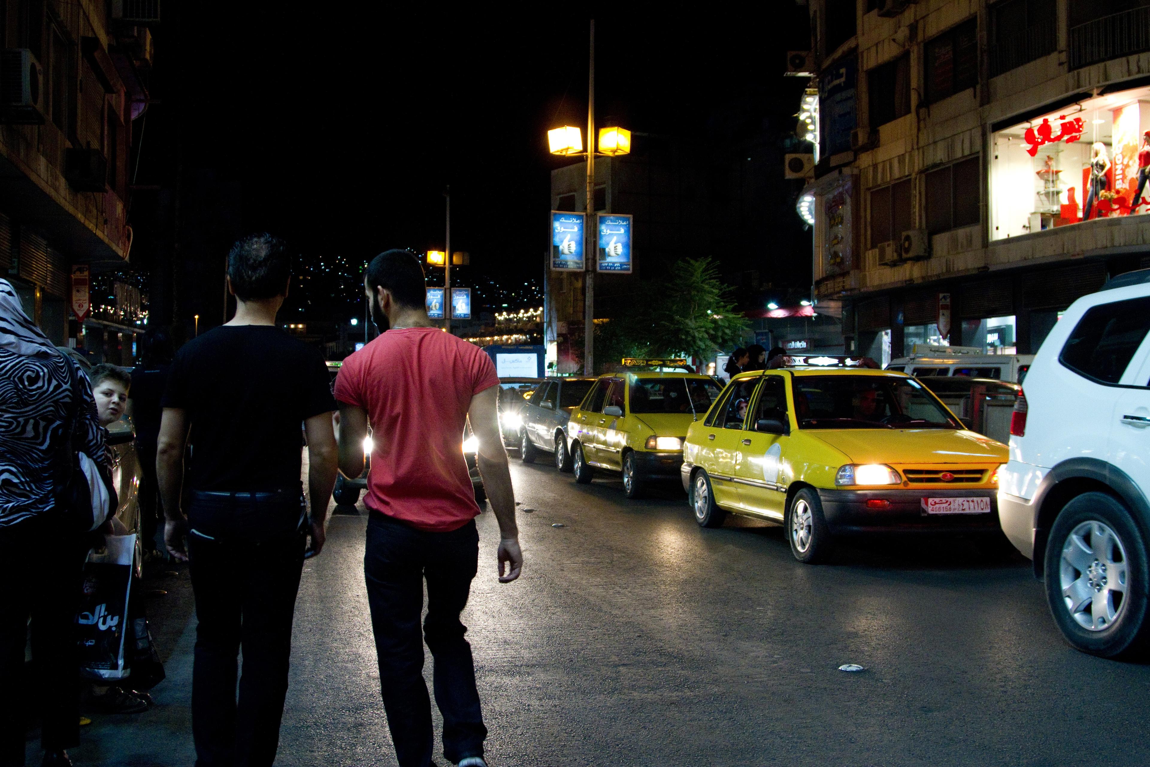 Photo of two men walking at night beside a street with yellow taxis and shops lit by streetlamps, with city lights in the background.