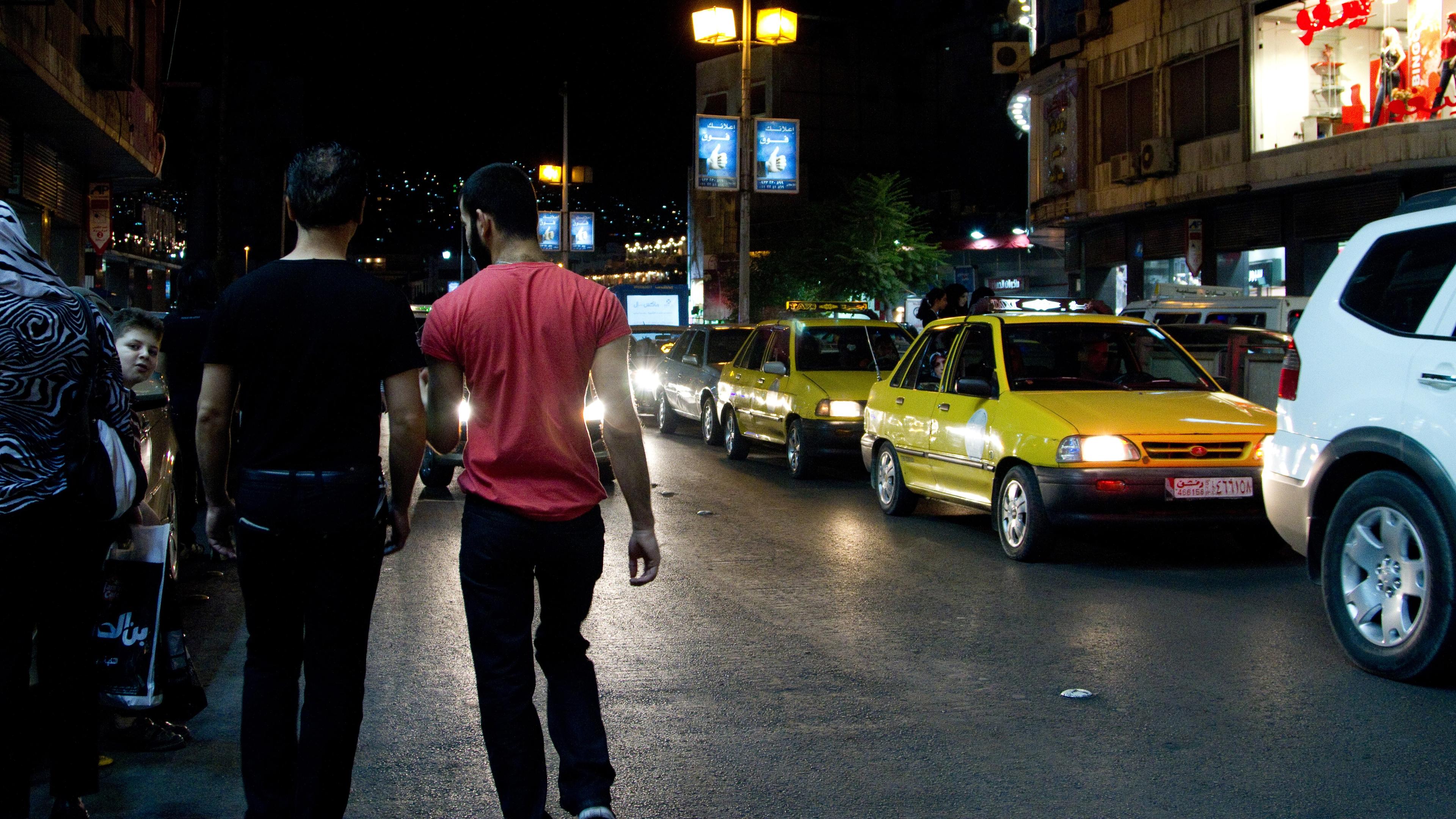 Photo of two men walking at night beside a street with yellow taxis and shops lit by streetlamps, with city lights in the background.