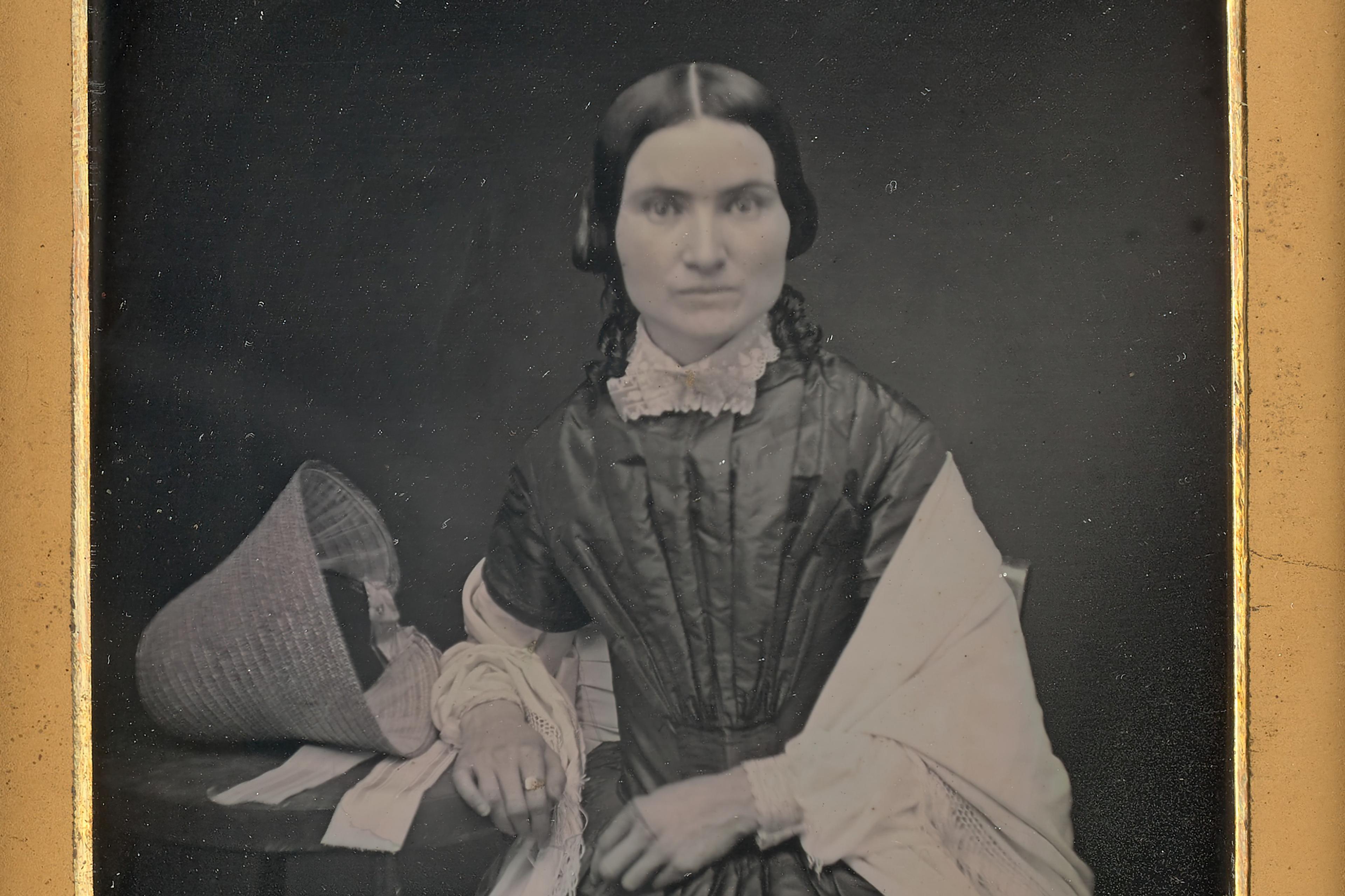 Daguerreotype portrait of a woman in 19th-century attire sitting with a hat on a table, framed in ornate gold and red.