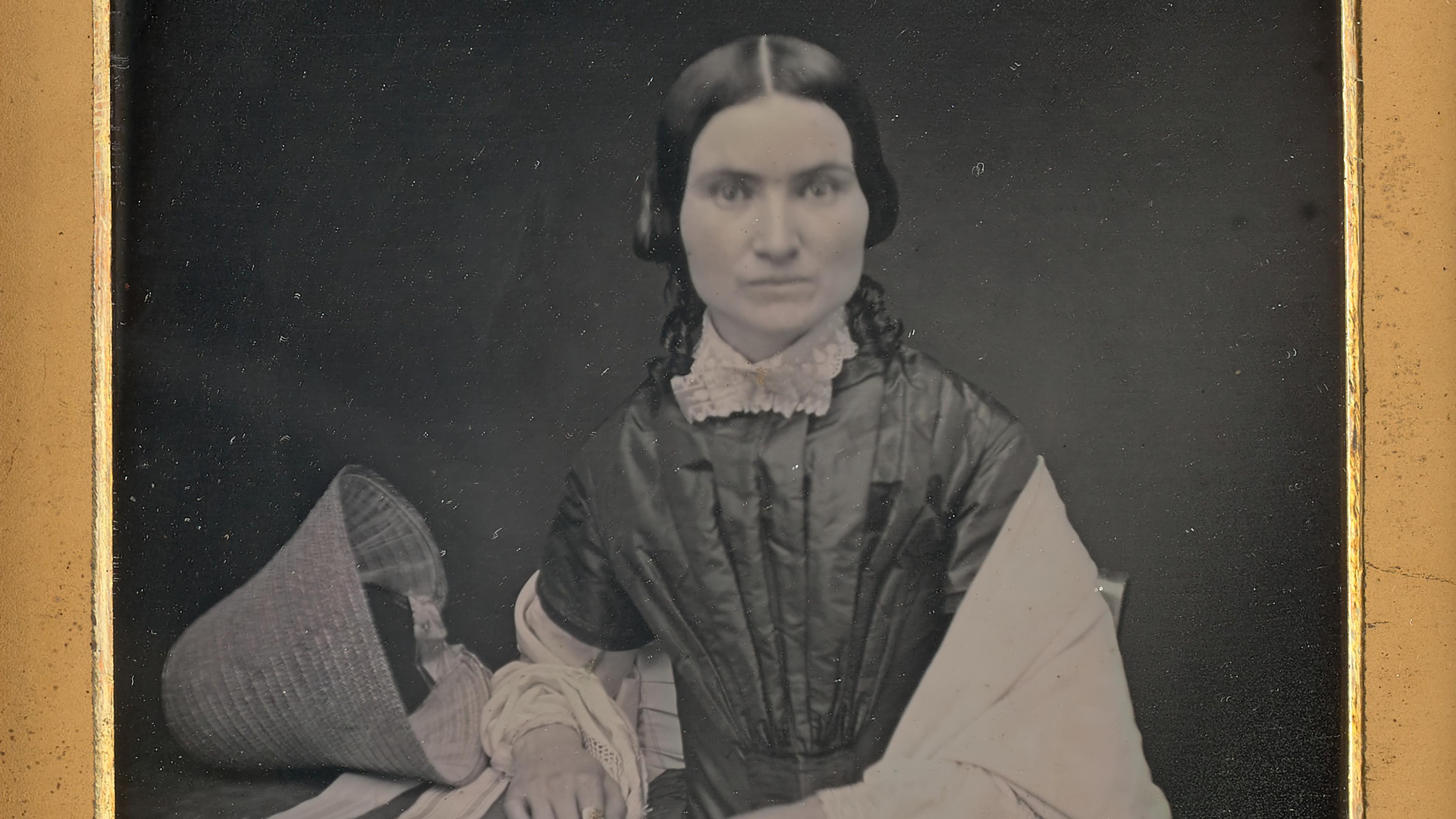 Daguerreotype portrait of a woman in 19th-century attire sitting with a hat on a table, framed in ornate gold and red.