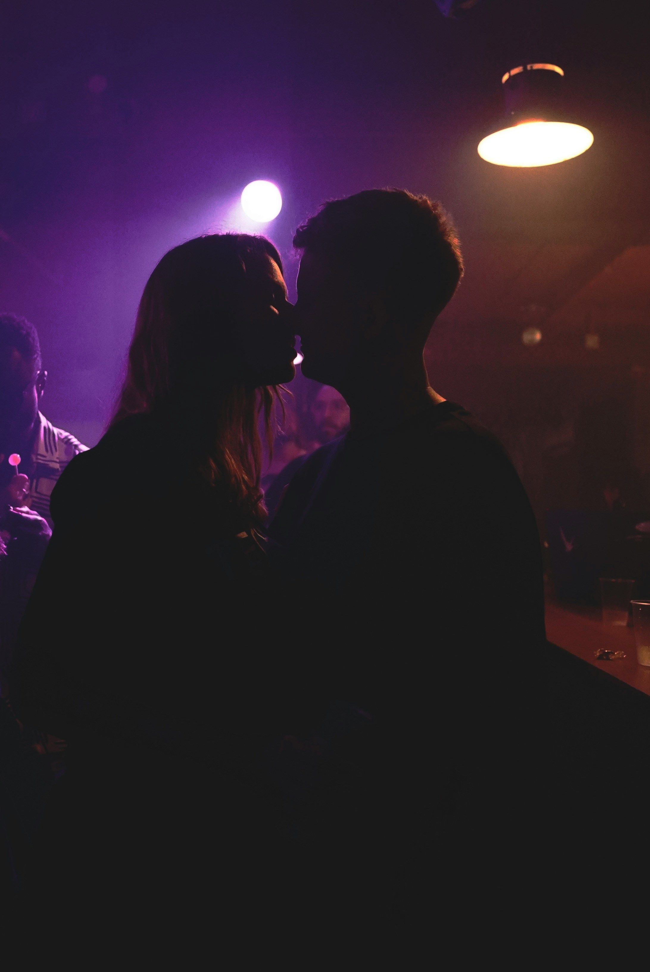 A silhouetted couple kissing in a dimly lit club with purple lighting and people in the background.