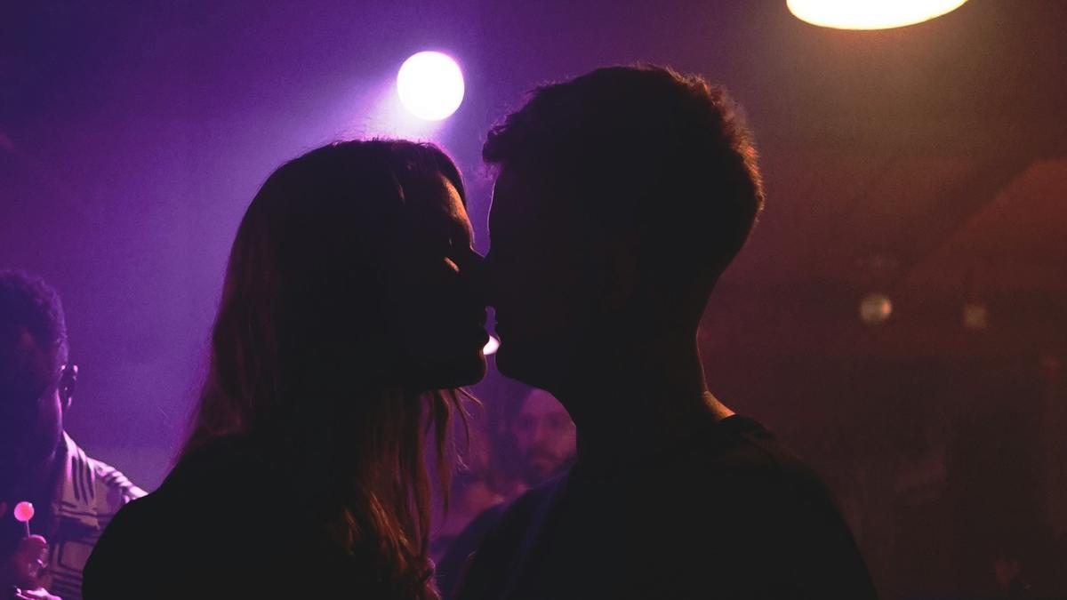 A silhouetted couple kissing in a dimly lit club with purple lighting and people in the background.