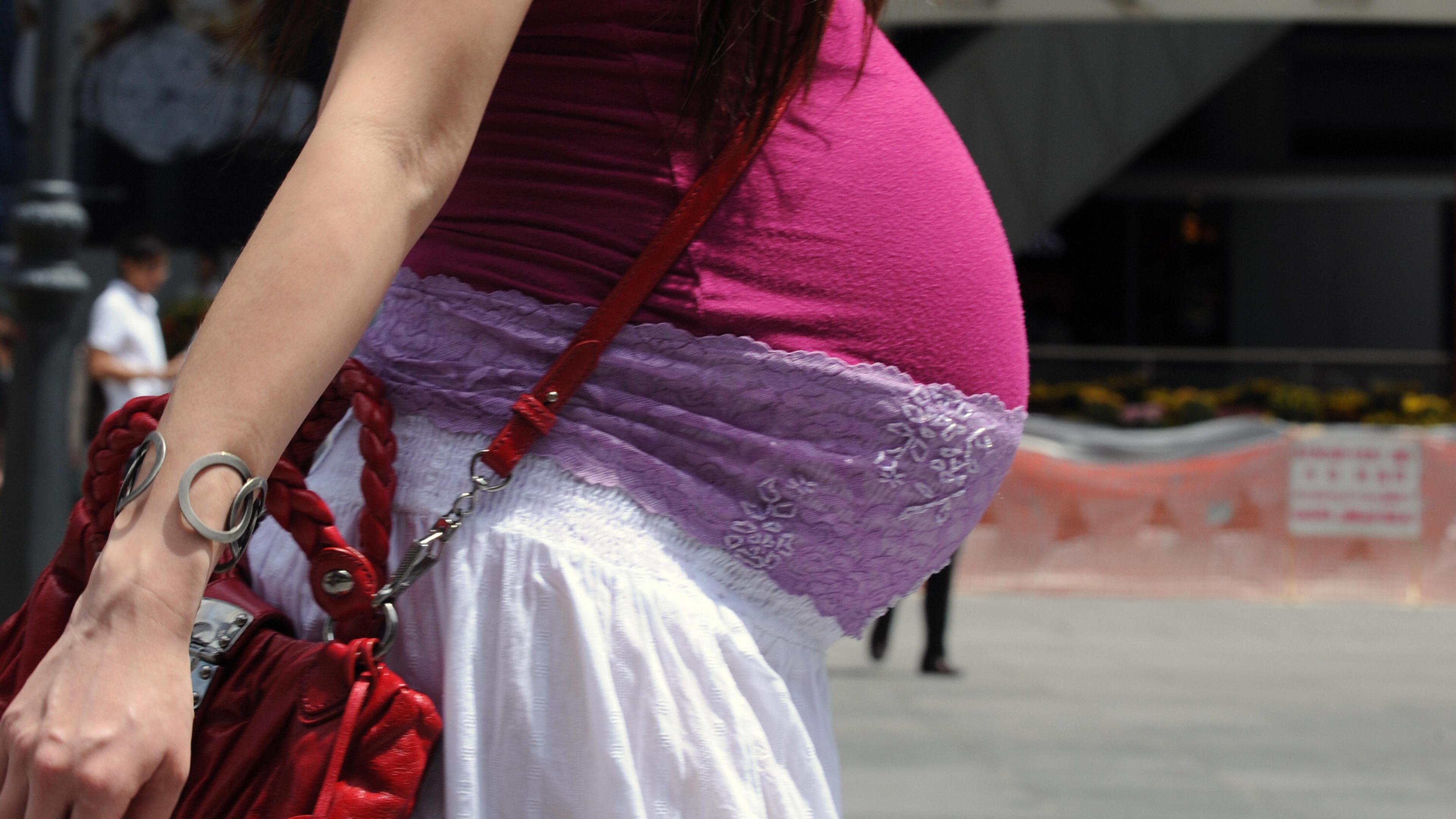 Photo of a pregnant woman in a pink vest and white skirt, holding a red bag, walking in an urban area.