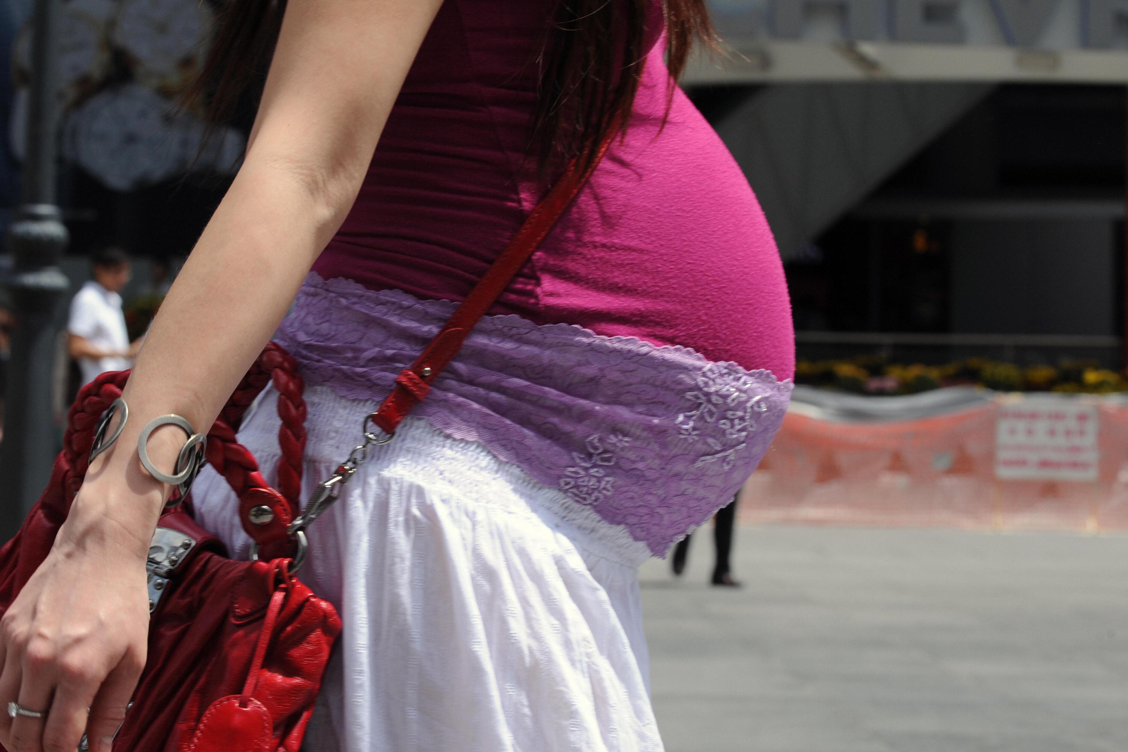 Photo of a pregnant woman in a pink vest and white skirt, holding a red bag, walking in an urban area.