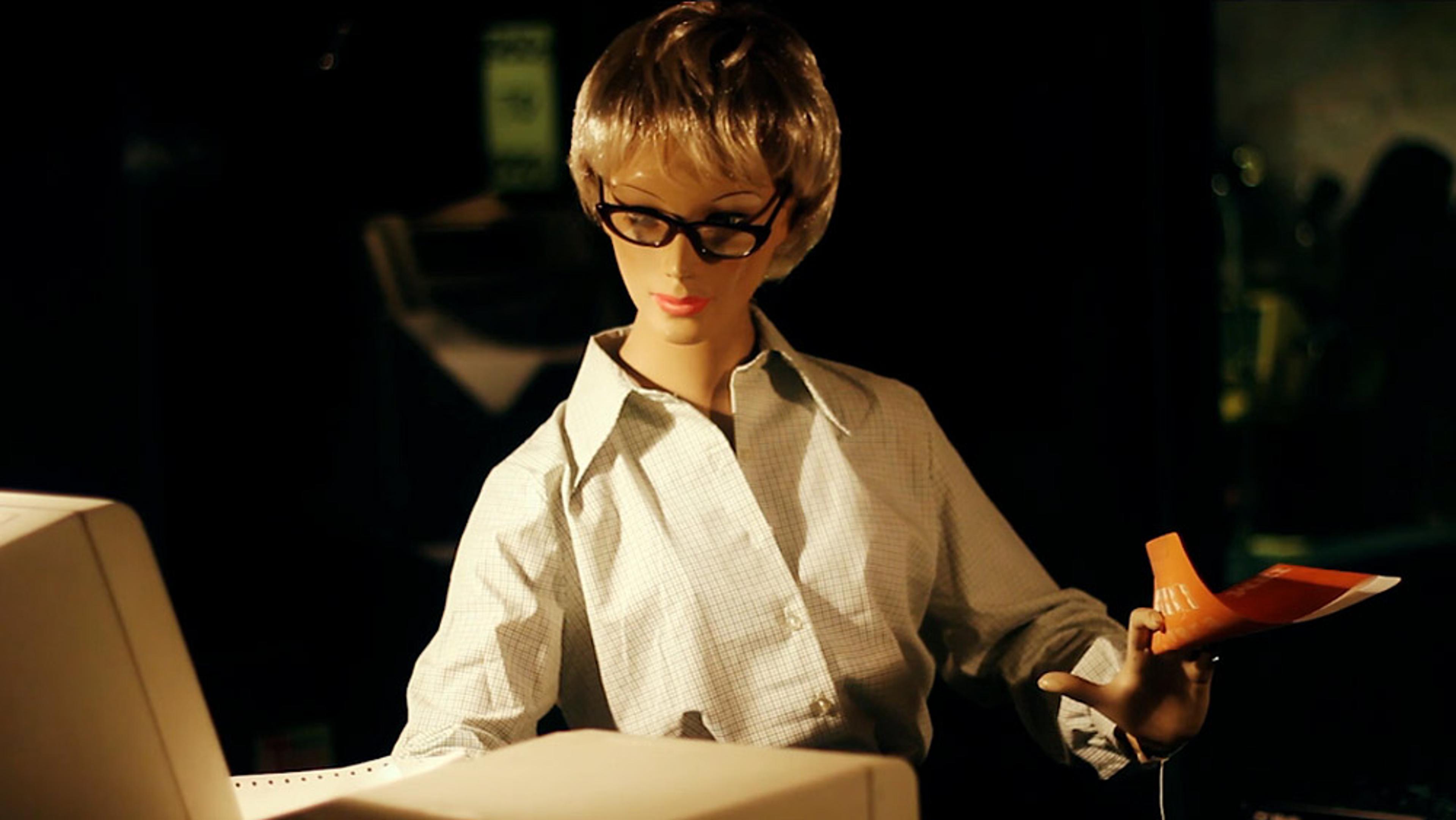 A mannequin with short hair and glasses in a dimly lit room, seated at a desk holding a booklet and typing.