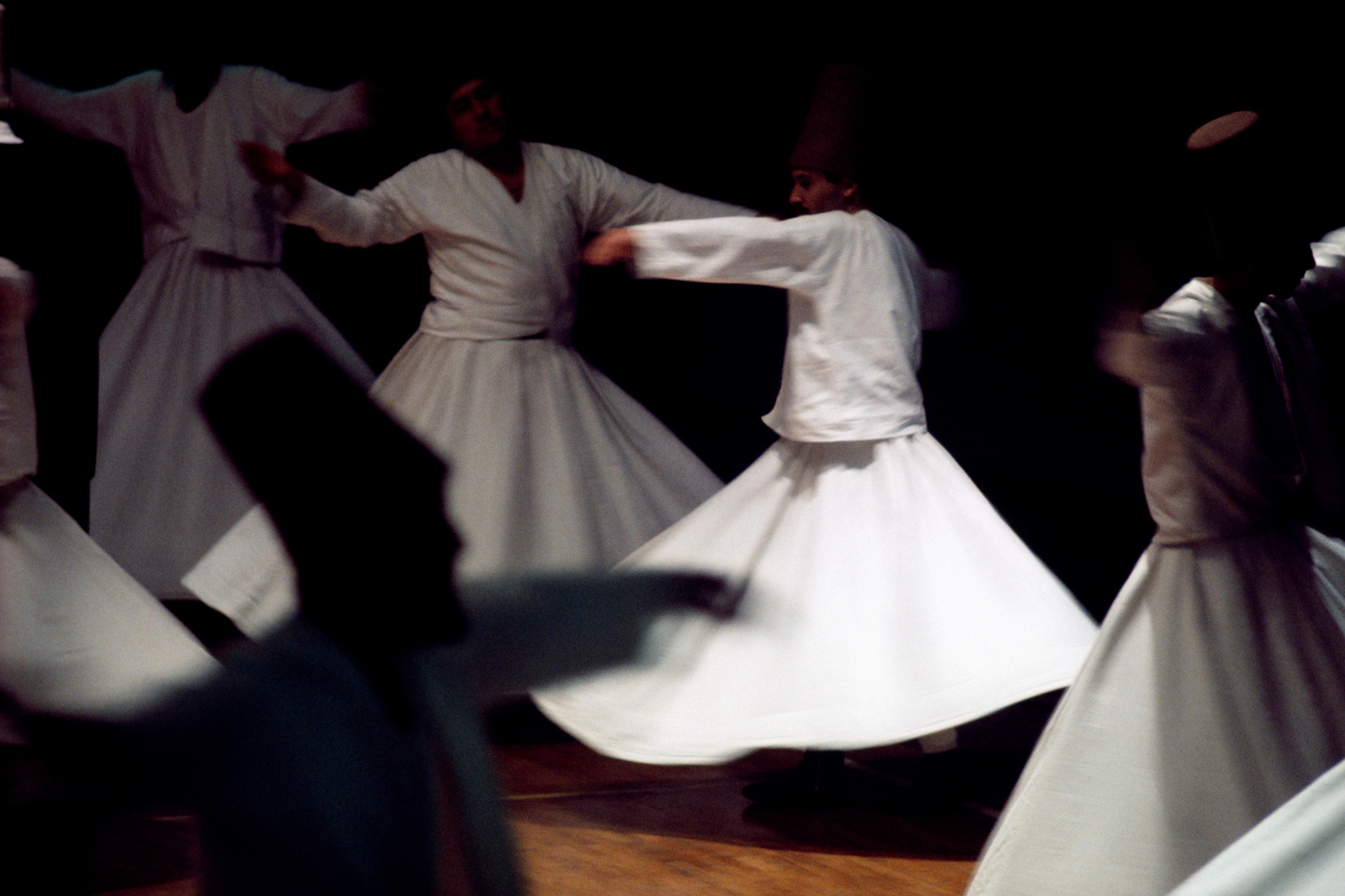 Photo of Sufi whirling dervishes in white robes spinning in a dimly lit room during a traditional dance performance.