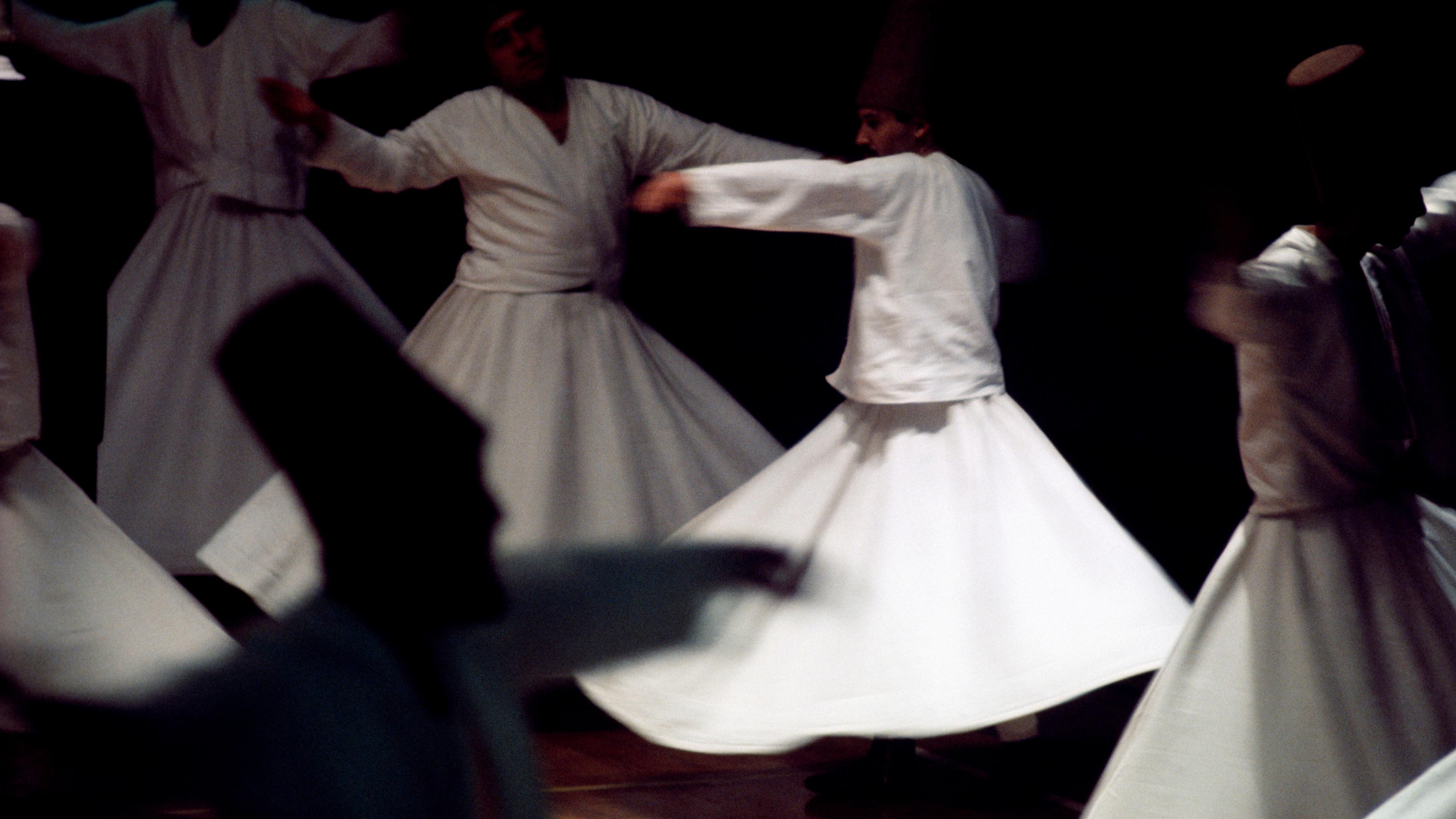 Photo of Sufi whirling dervishes in white robes spinning in a dimly lit room during a traditional dance performance.