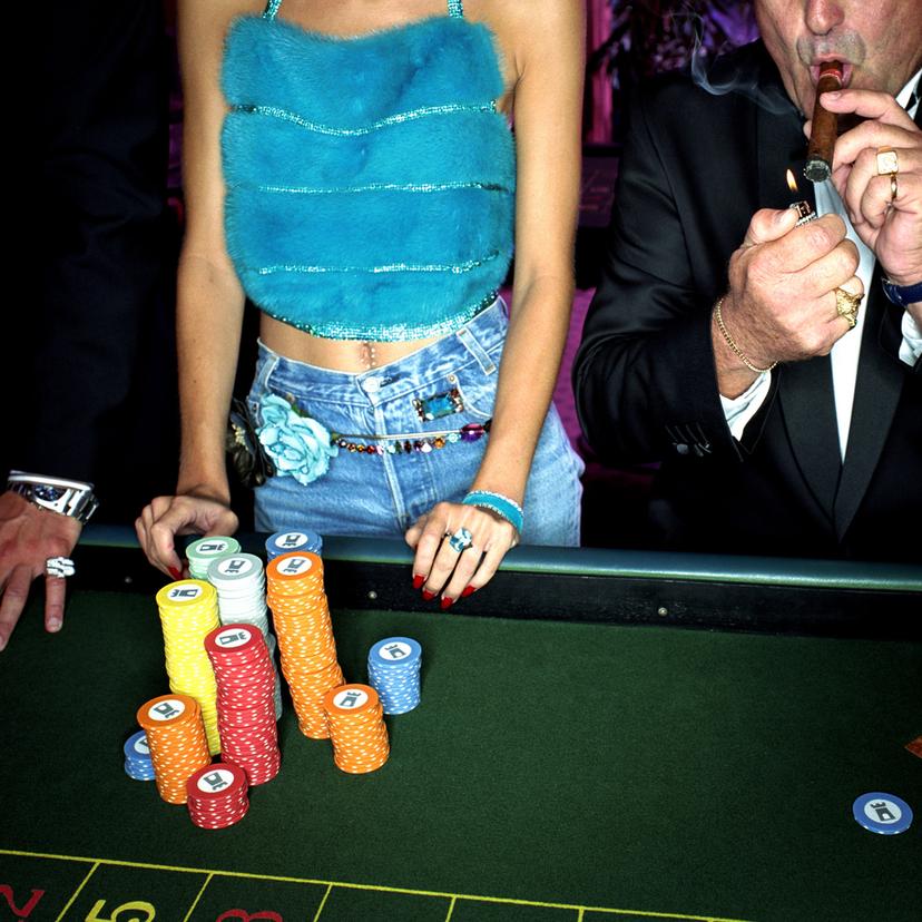 A young woman and an older man at a poker table with colourful chips, the man wears a suit and lights a cigar, the woman wears jeans and a fluffy turquoise top.