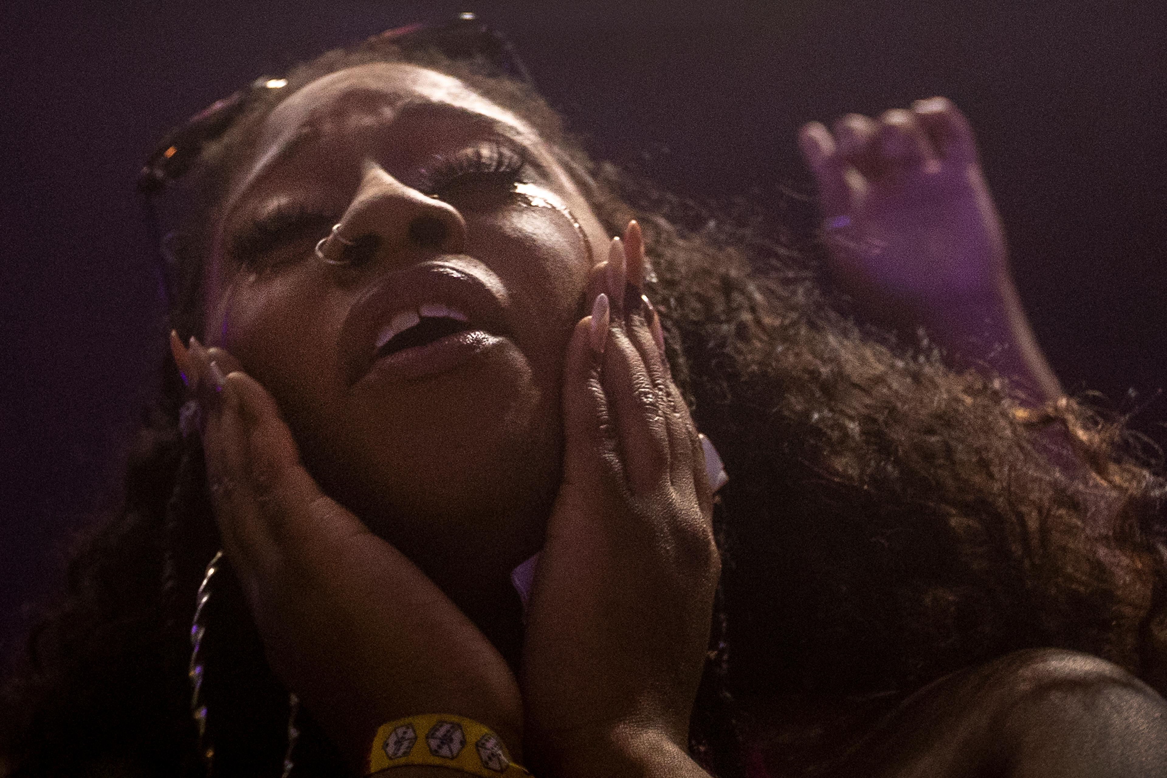 Photo of a person with eyes closed, hands on cheeks, wearing a wristband and large hoop earrings in a dimly lit setting.
