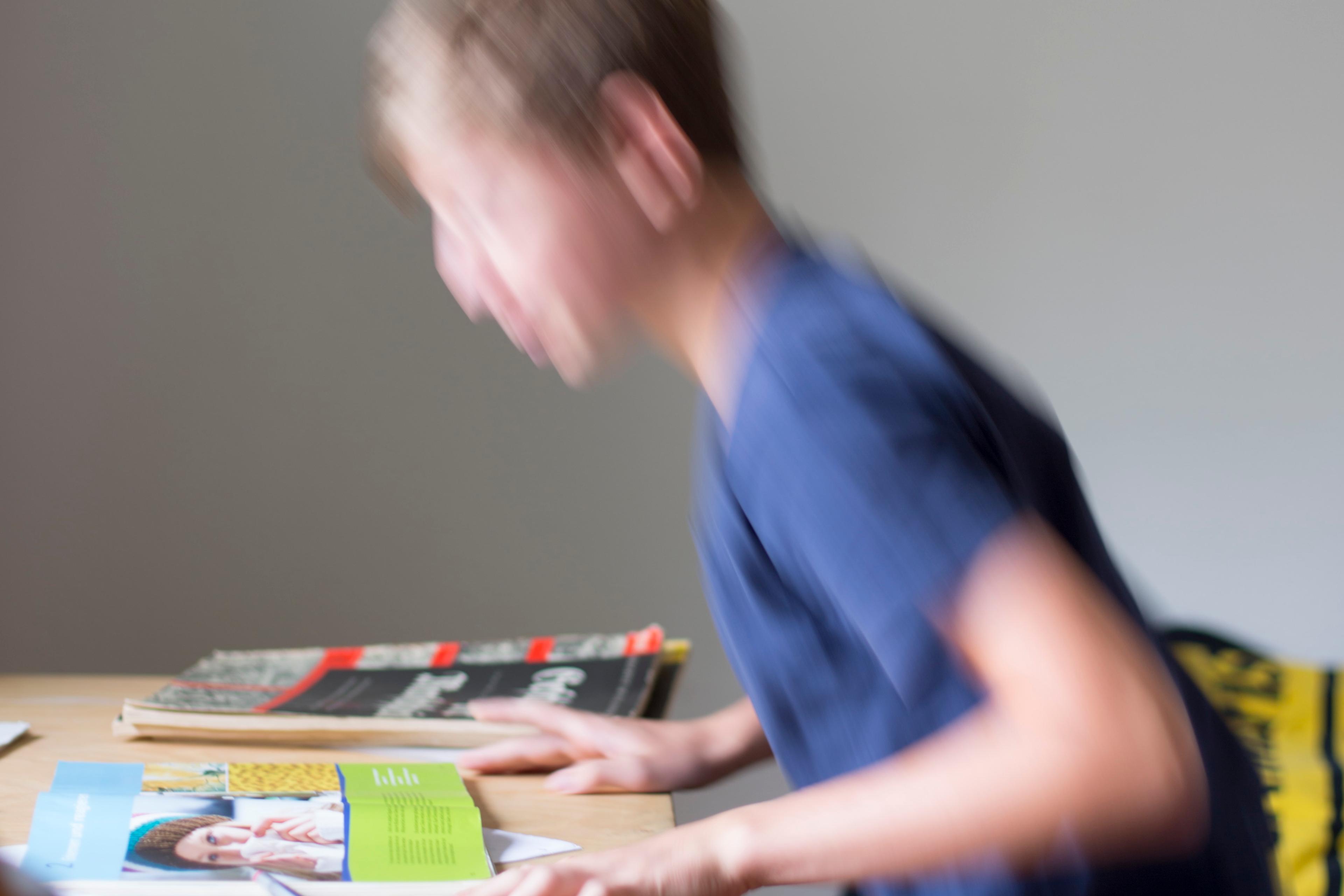 Blurry photo of a child in a blue shirt sitting at a desk with open books and magazines.