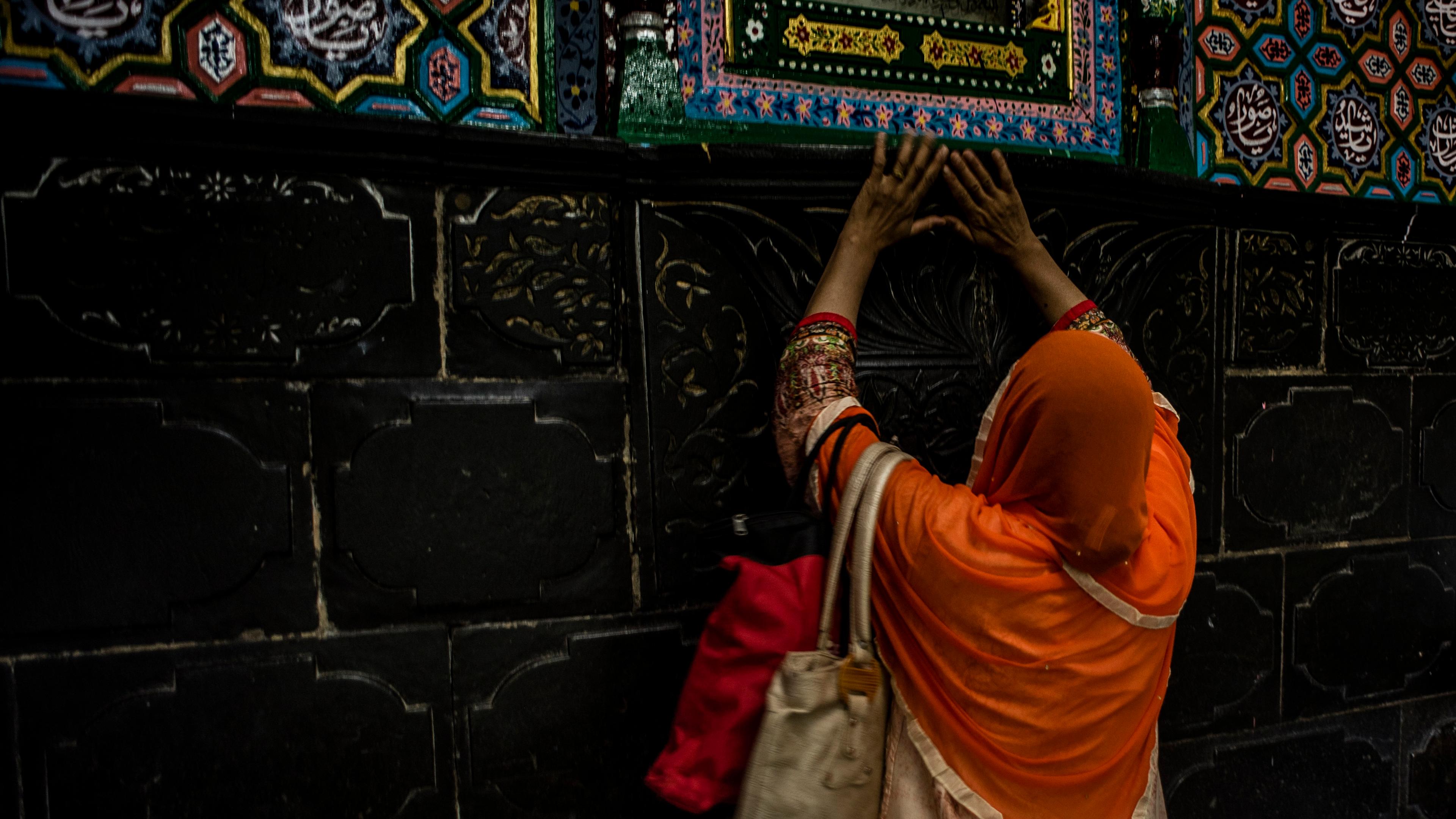 Photo of a woman in an orange shawl touching a decorative wall with colourful, intricate patterns and Islamic calligraphy.