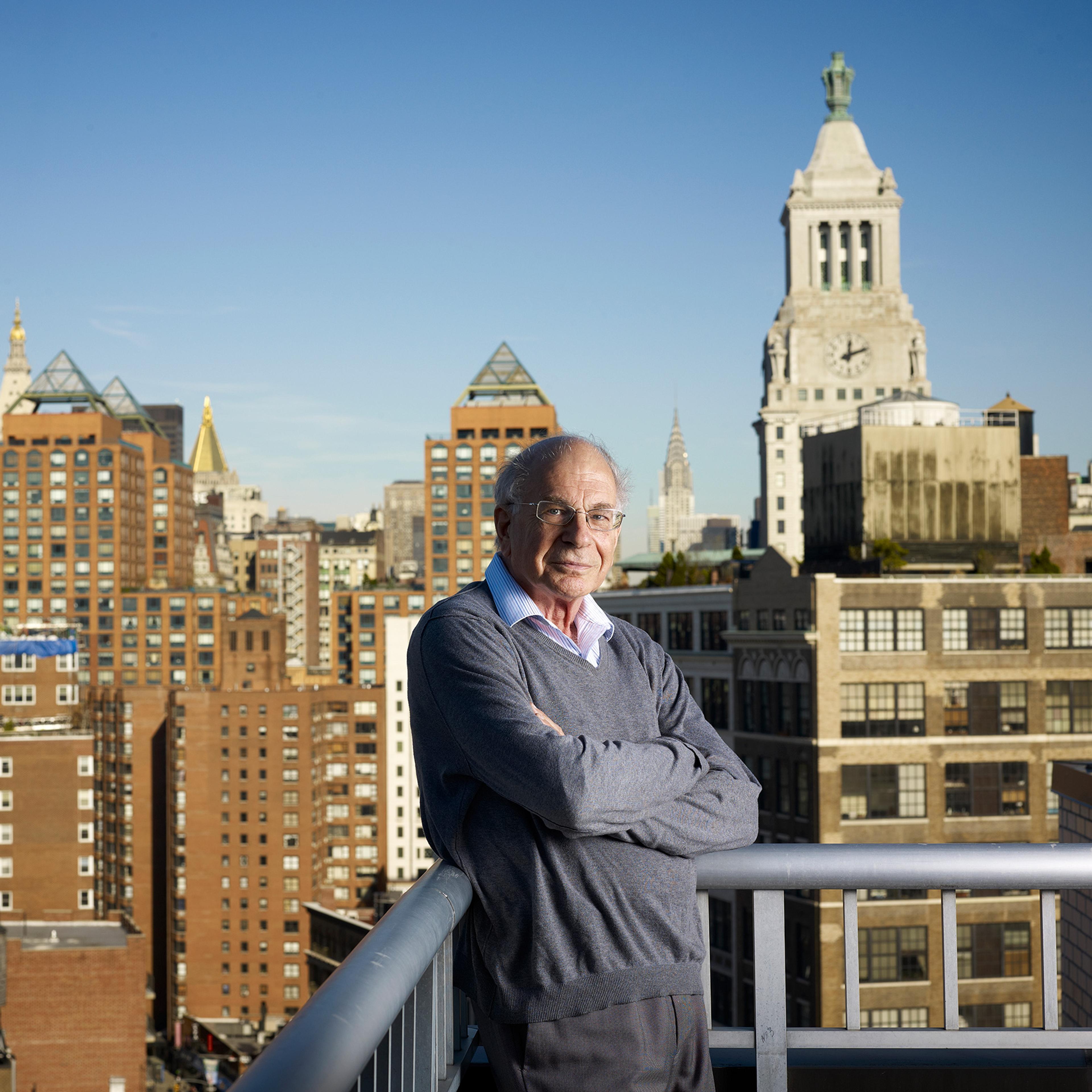 Daniel Kahneman standing on a balcony, arms crossed, with New York City skyline and buildings in the background under a clear blue sky.