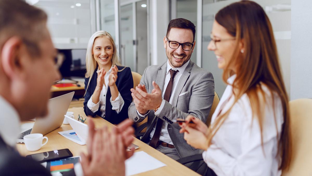 A group of office workers turned to face a colleague, clapping and smiling, in a modern meeting room setting.