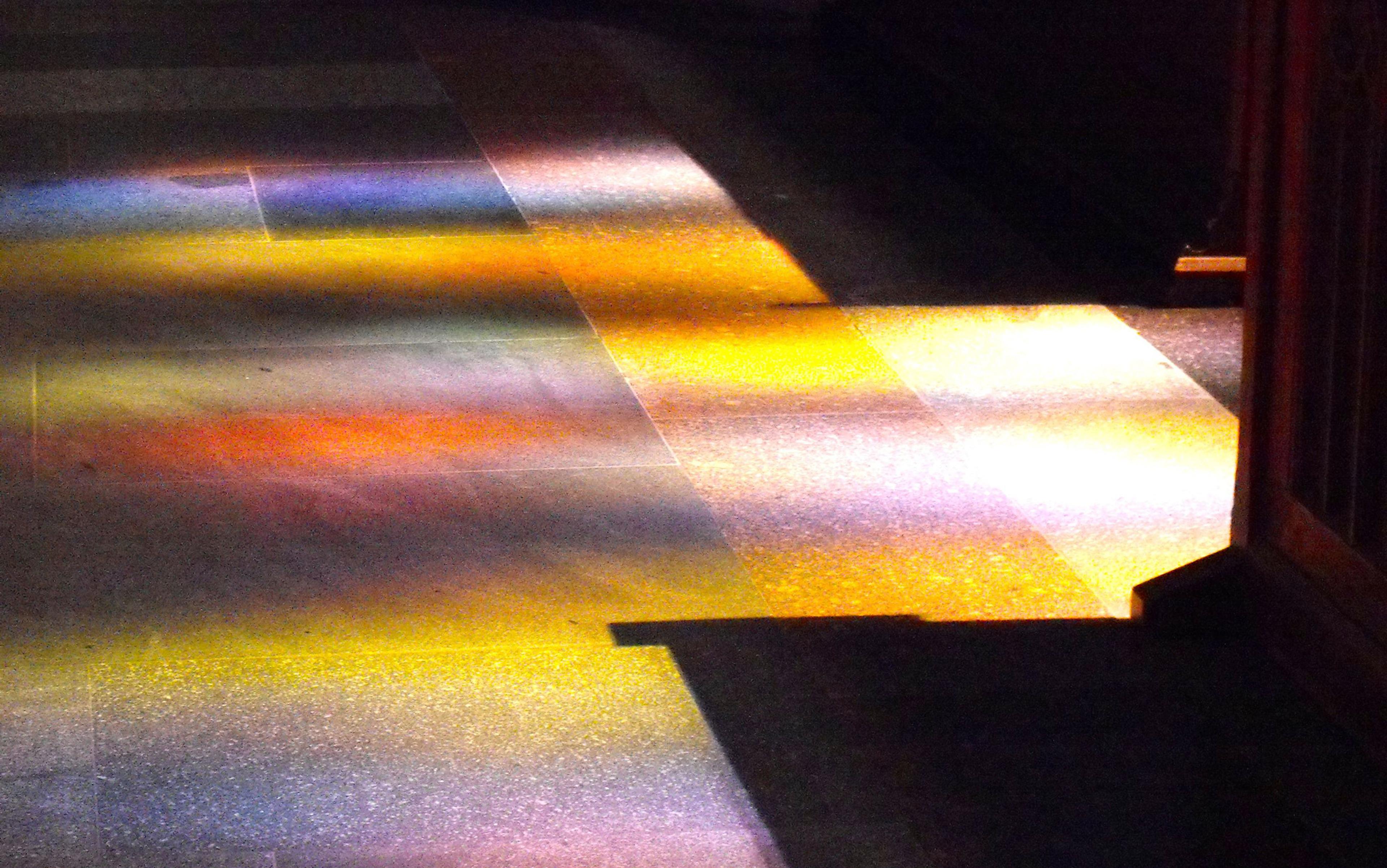 Photo of colourful light reflections on a dark tiled floor, possibly from a stained glass window in a dimly lit interior space.