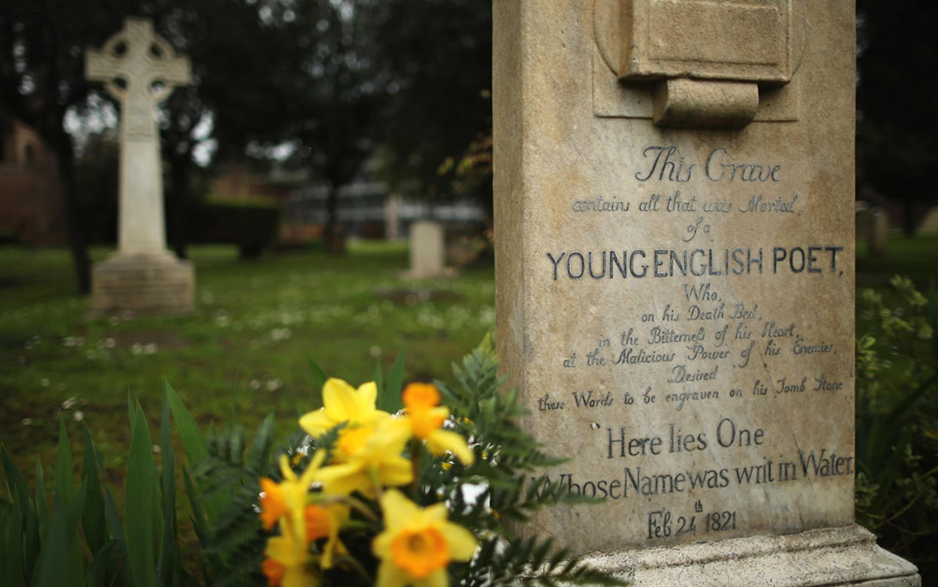 A gravestone inscribed “Young English Poet” with daffodils in the foreground and a Celtic cross in a cemetery in the background.