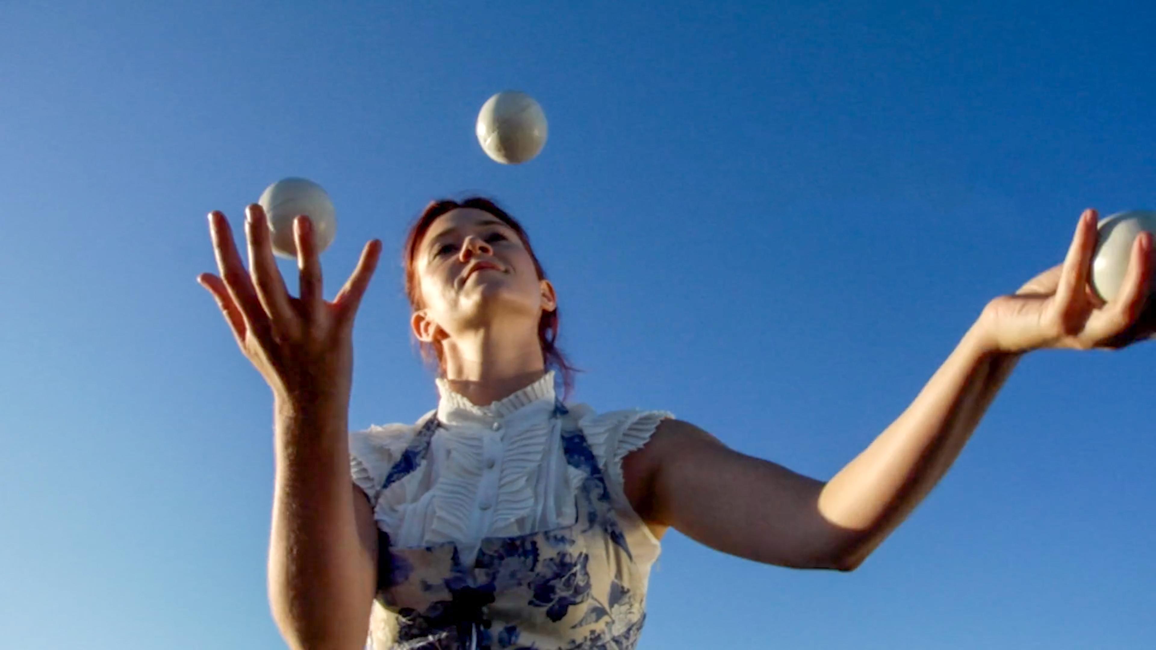 Photo of a woman juggling three white balls against a clear blue sky, wearing a white ruffled blouse and floral apron.