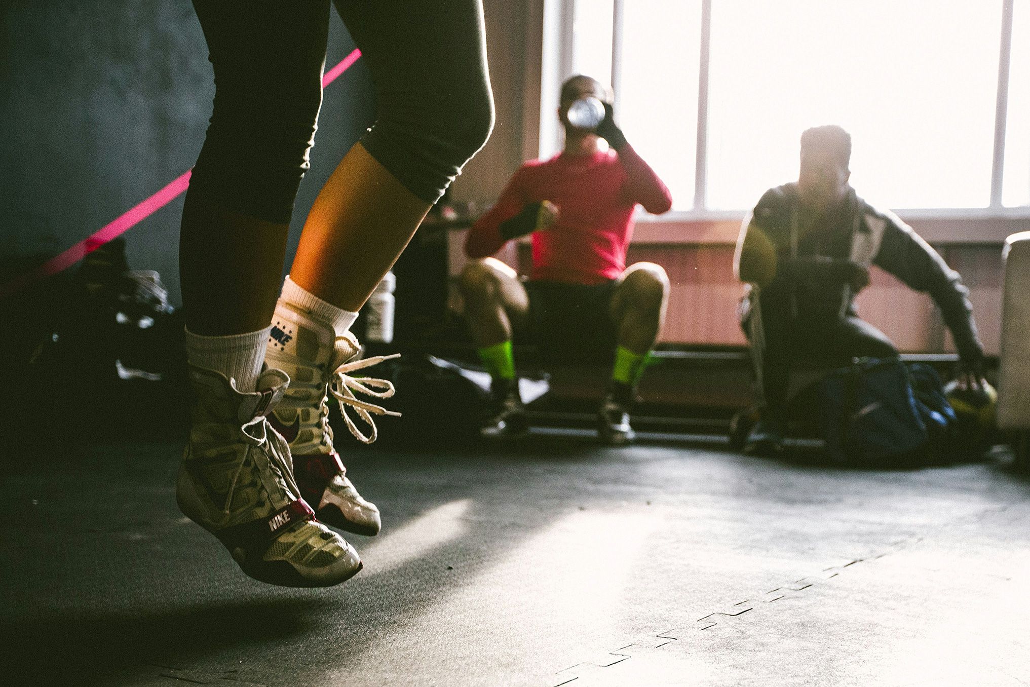 Photo of person skipping in training shoes indoors, with two others resting in the background near a window.