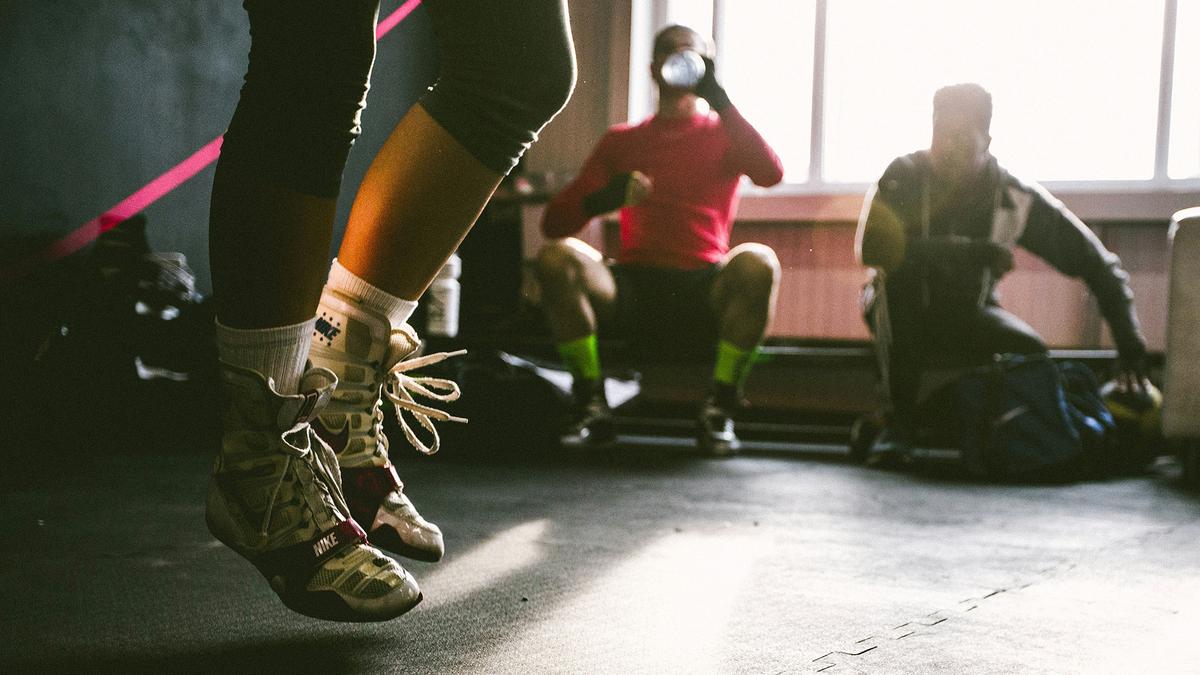 Photo of person skipping in training shoes indoors, with two others resting in the background near a window.