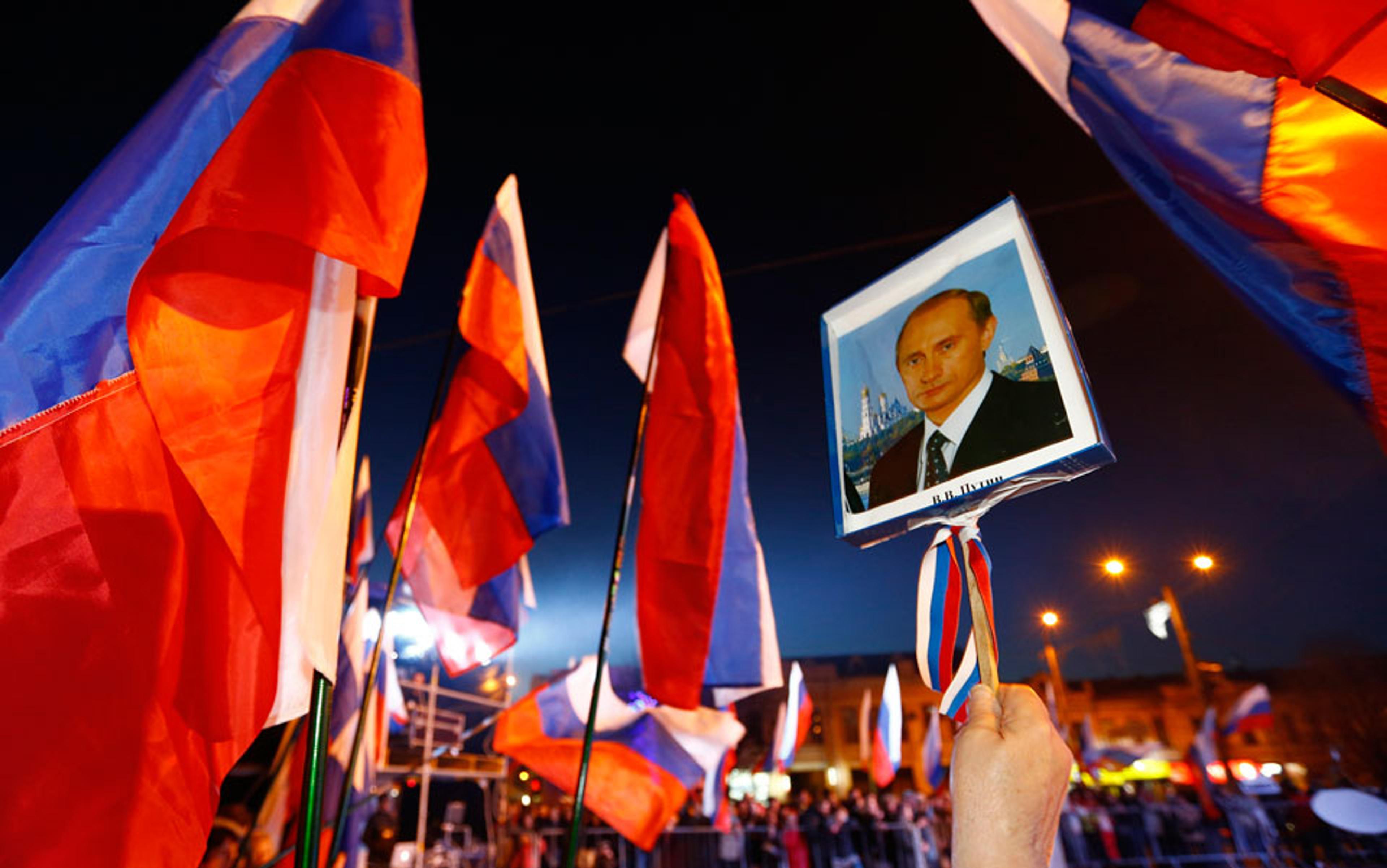 A person holding a placard with a portrait of Putin amid many Russian flags at a night-time outdoor event with street lights.