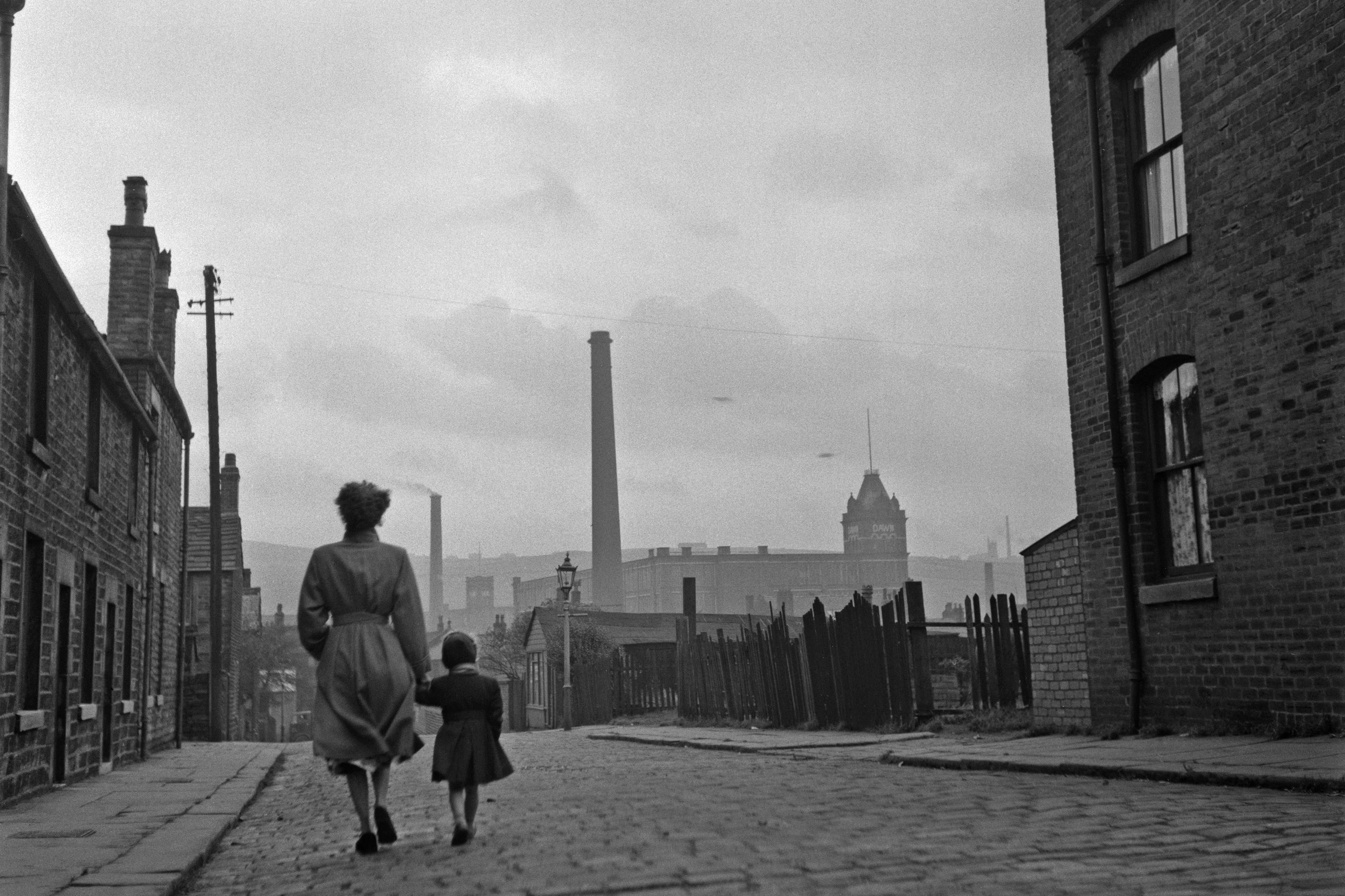 Black and white photo of a woman and child walking on a cobbled street in an industrial town with chimneys in the background.