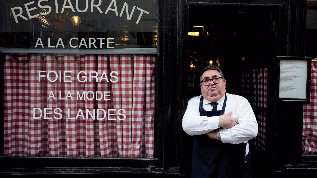 A man with crossed arms and a serious expression wearing an apron stands in the doorway of a restaurant with checkered curtains and menu on display.
