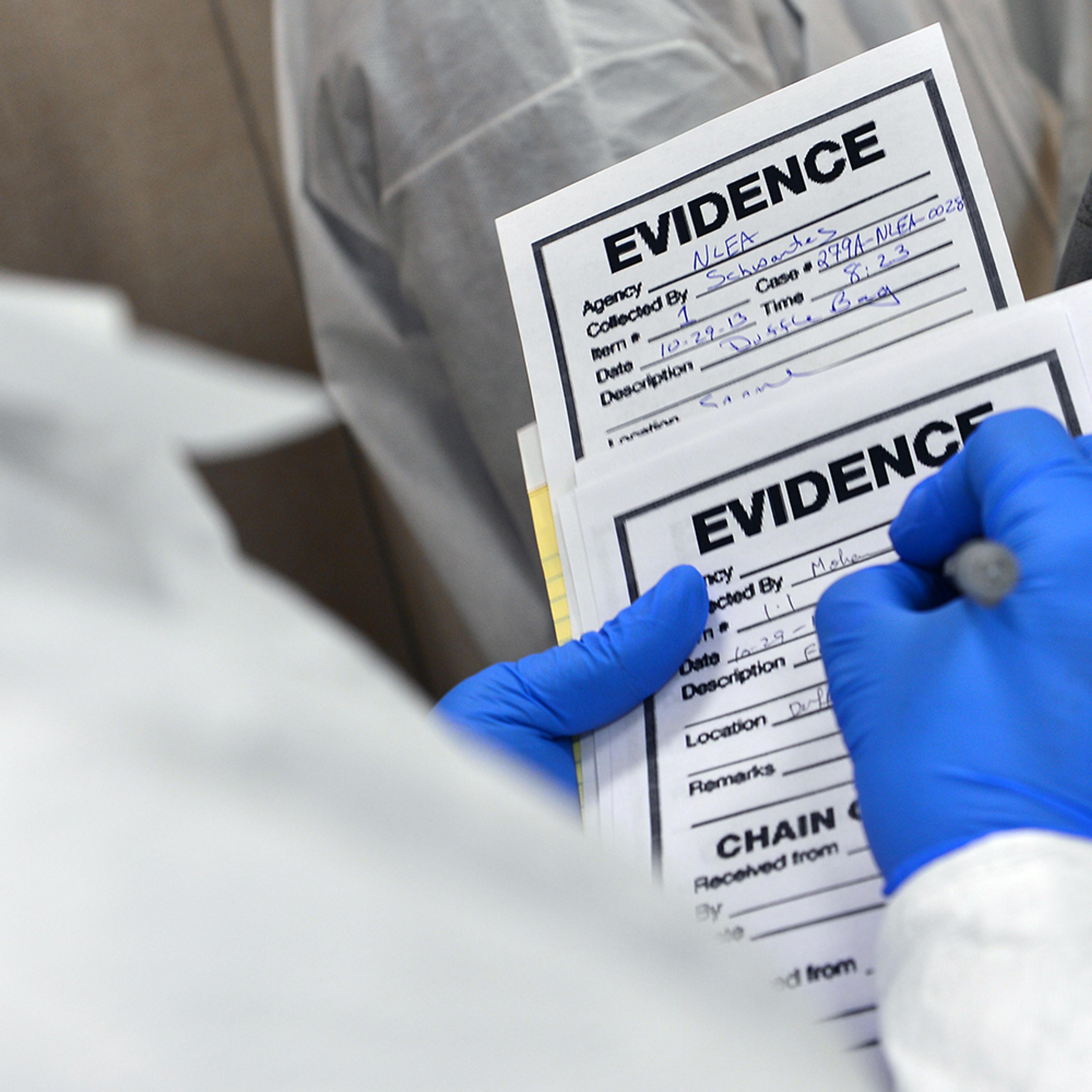 A person in protective gear writing on an evidence form with a pen, wearing blue gloves and holding two evidence forms.