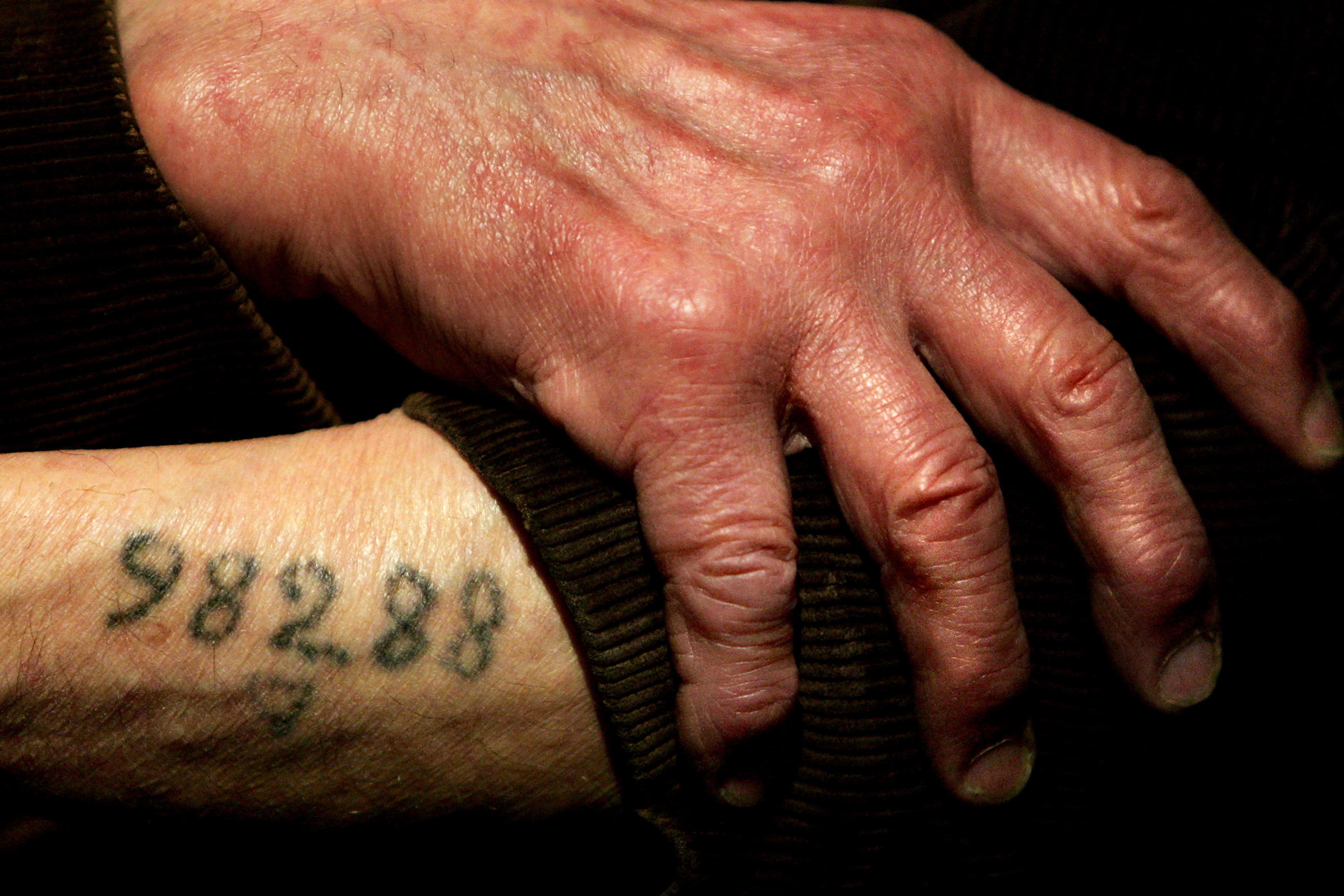 Photo of an elderly hand resting on a wrist, tattooed with the number 58288, on a dark fabric background.